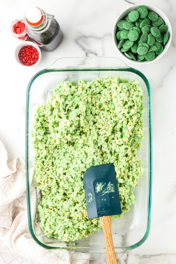 Spatula smoothing out green dyed Rice Krispies bars in glass baking dish