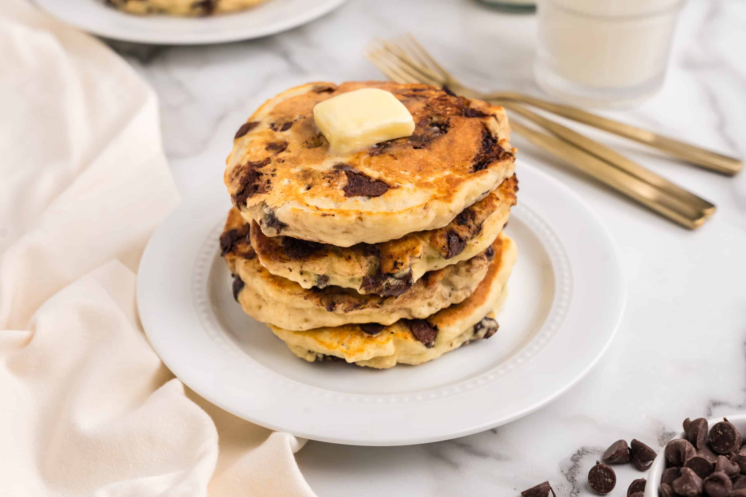 A stack of pancakes displayed on a white plate with silverware on the table beside the white plate.