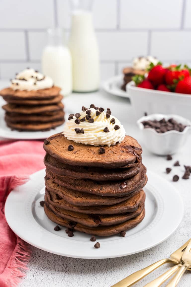 Chocolate Pancakes stacked on white plate topped with whipped cream and chocolate chips