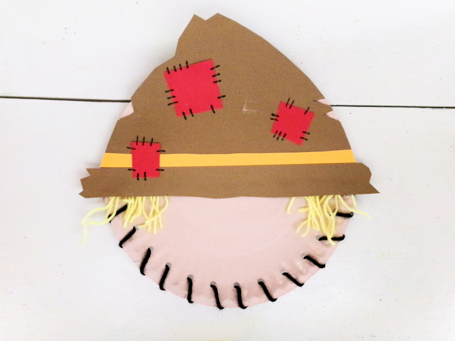Gluing the hat with added accessories to the paper plate face for the Paper Plate Scarecrow Craft