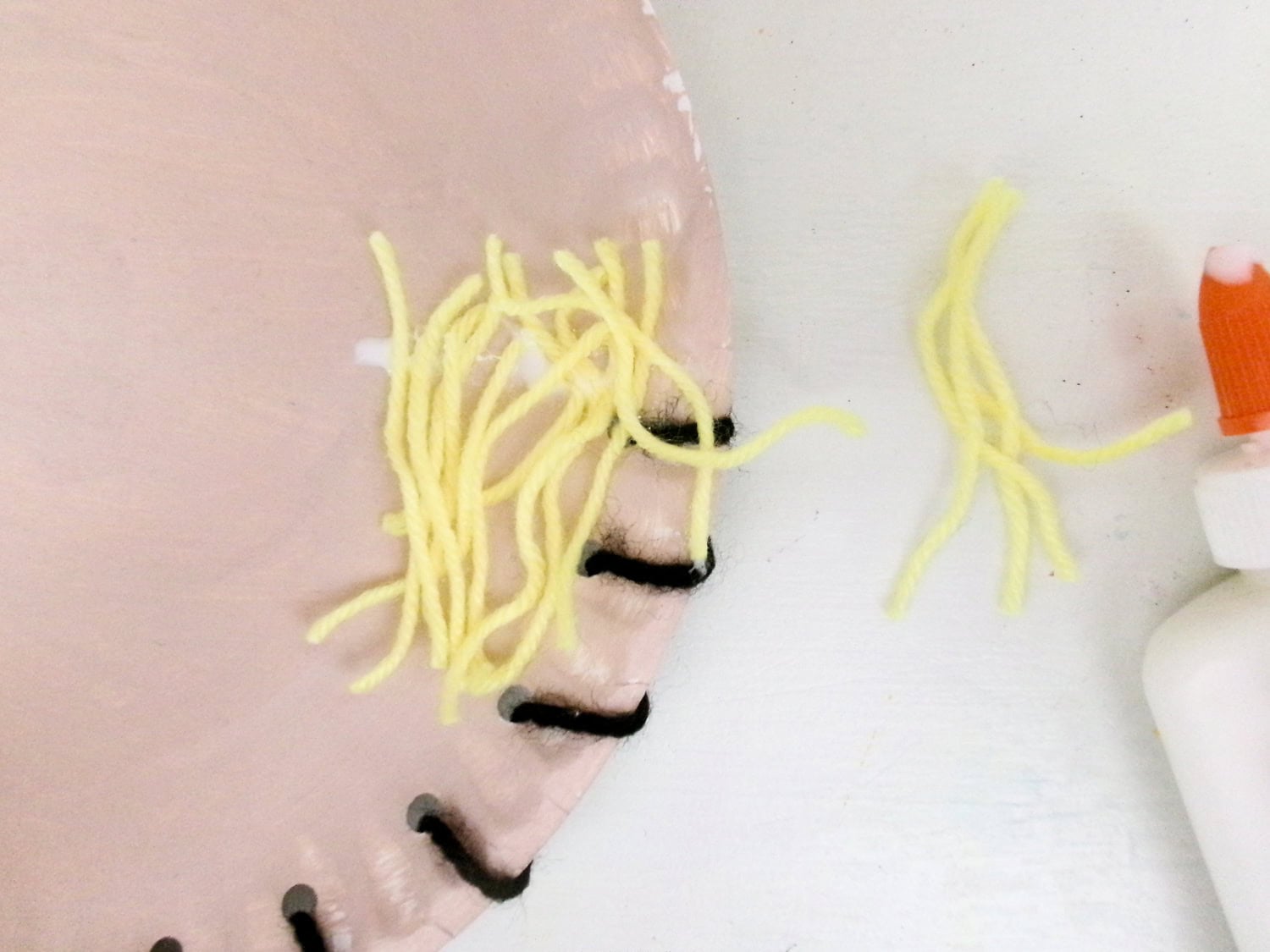 Gluing yellow yarn pieces to the painted face just above the threaded black yarn for the Paper Plate Scarecrow Craft