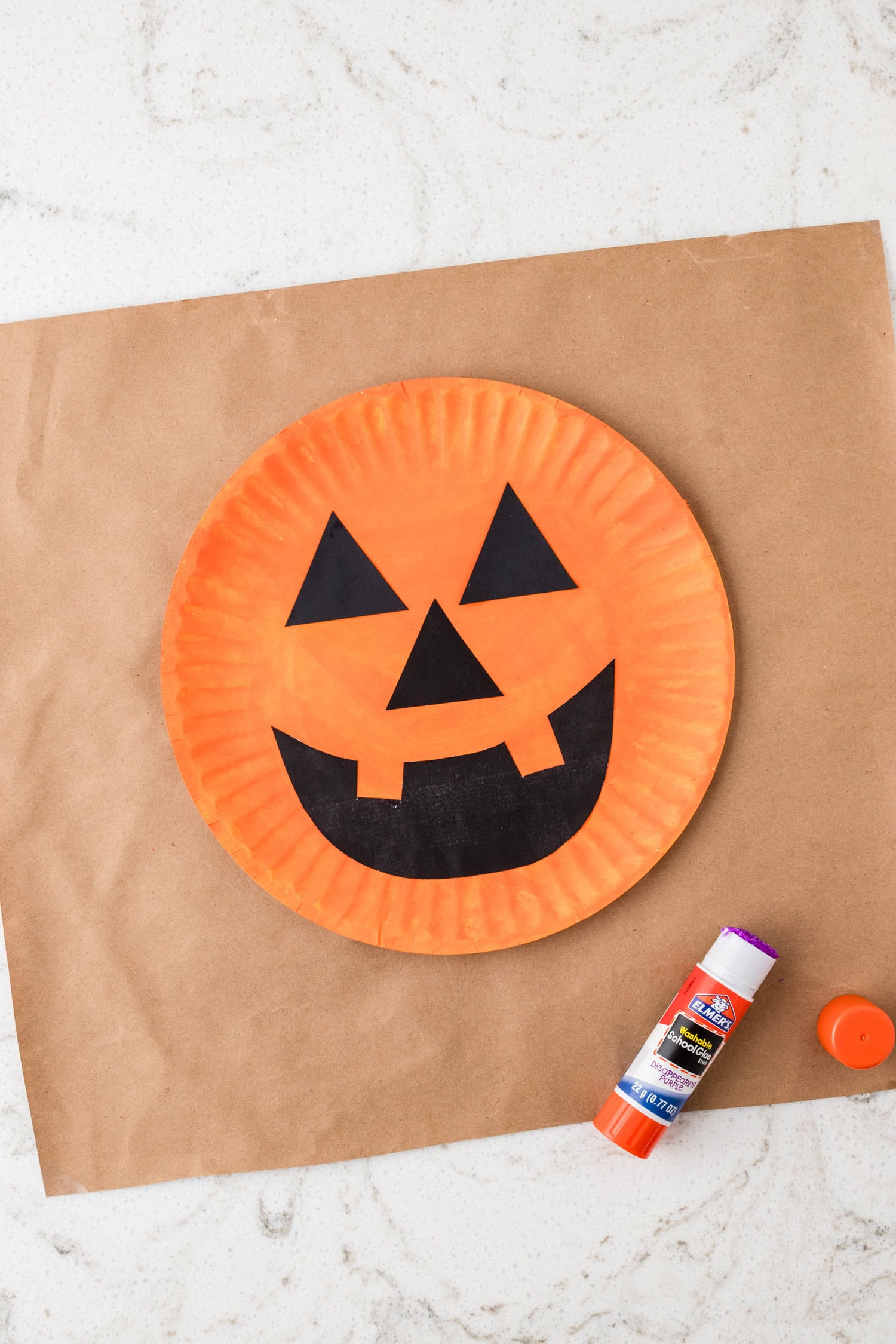 Using a glue stick to adhere the pumpkin face to the painted paper plate for the Paper Plate Pumpkin Craft