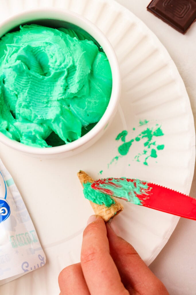 Overhead picture of red plastic knife spreading green frosting onto bugle.