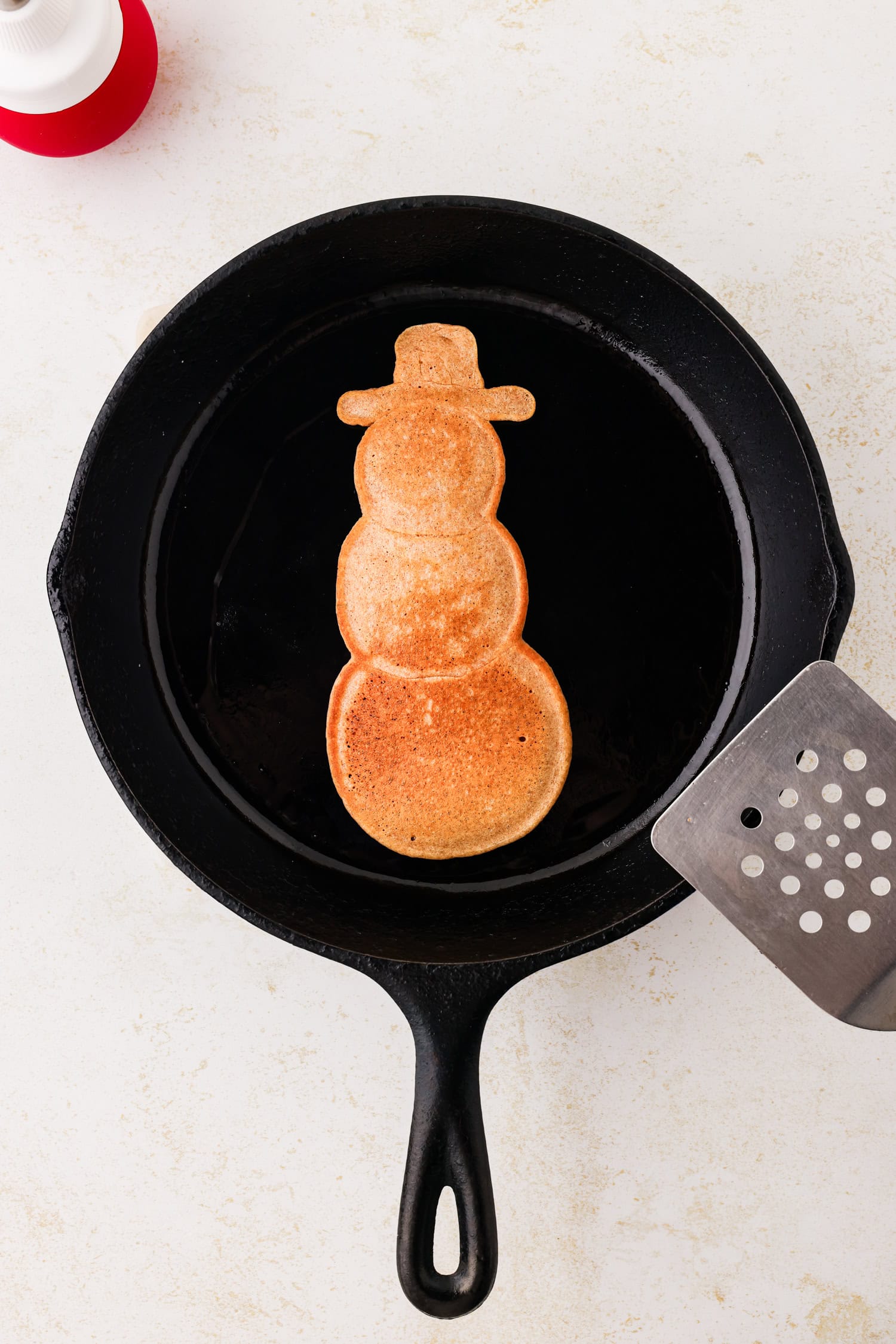 Snowman pancake cooked on a skillet with a metal spatula next to it.
