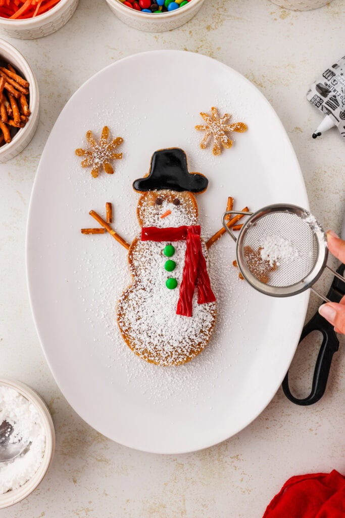 Completed snowman pancake with sifter pouring powdered sugar on it, on an white oval plate.