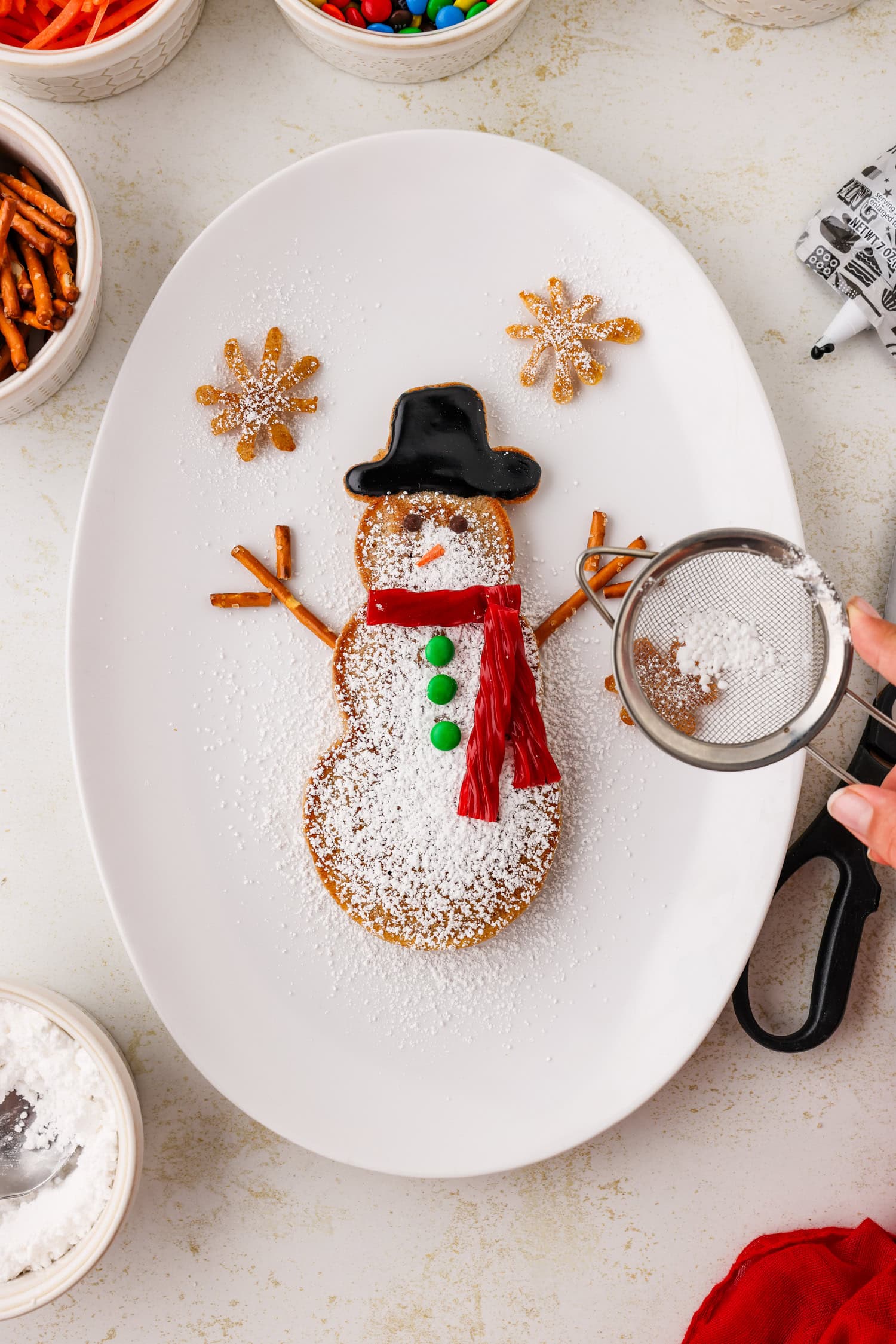 Completed snowman pancake with sifter pouring powdered sugar on it, on an white oval plate.