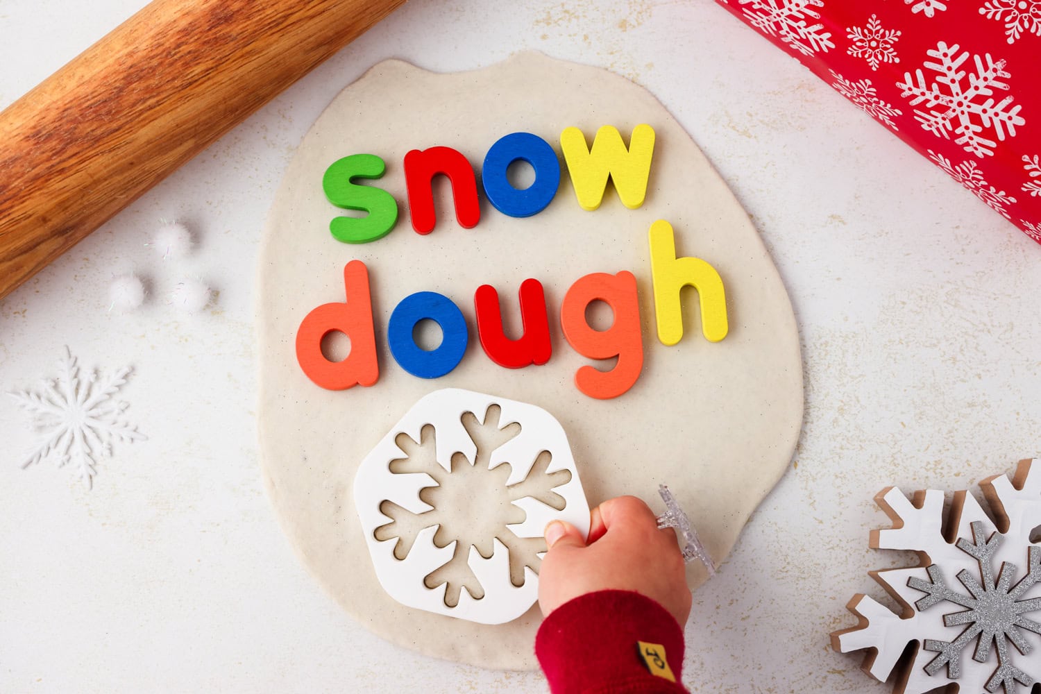 Overhead picture of snow dough with letters spelling out snow dough and a hand placing a cookie cutter in it.
