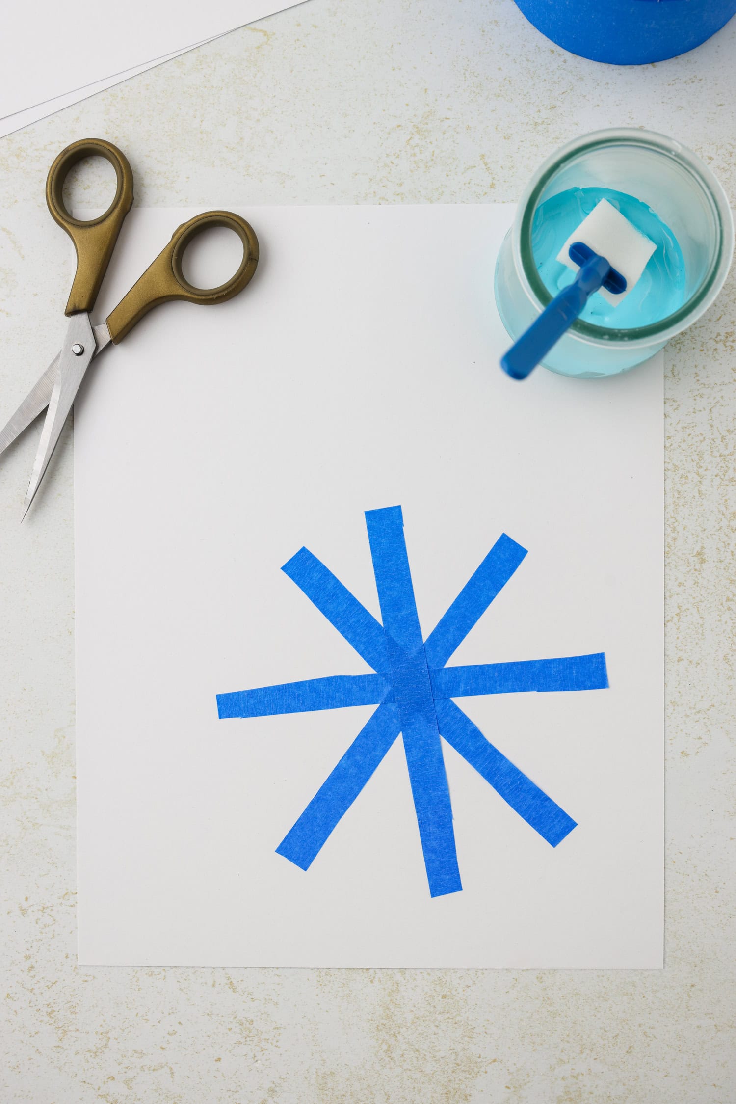 Overhead picture of blue painters tape on cardstock paper in pattern of a snowflake.