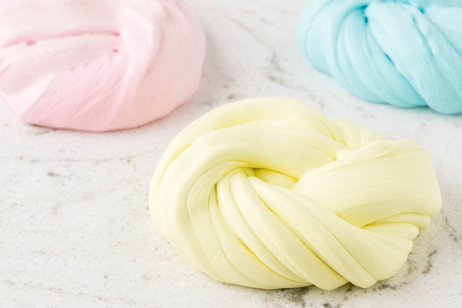 Three swirled piles of pastel-colored butter slime in yellow, pink, and blue, placed on a white marble surface.