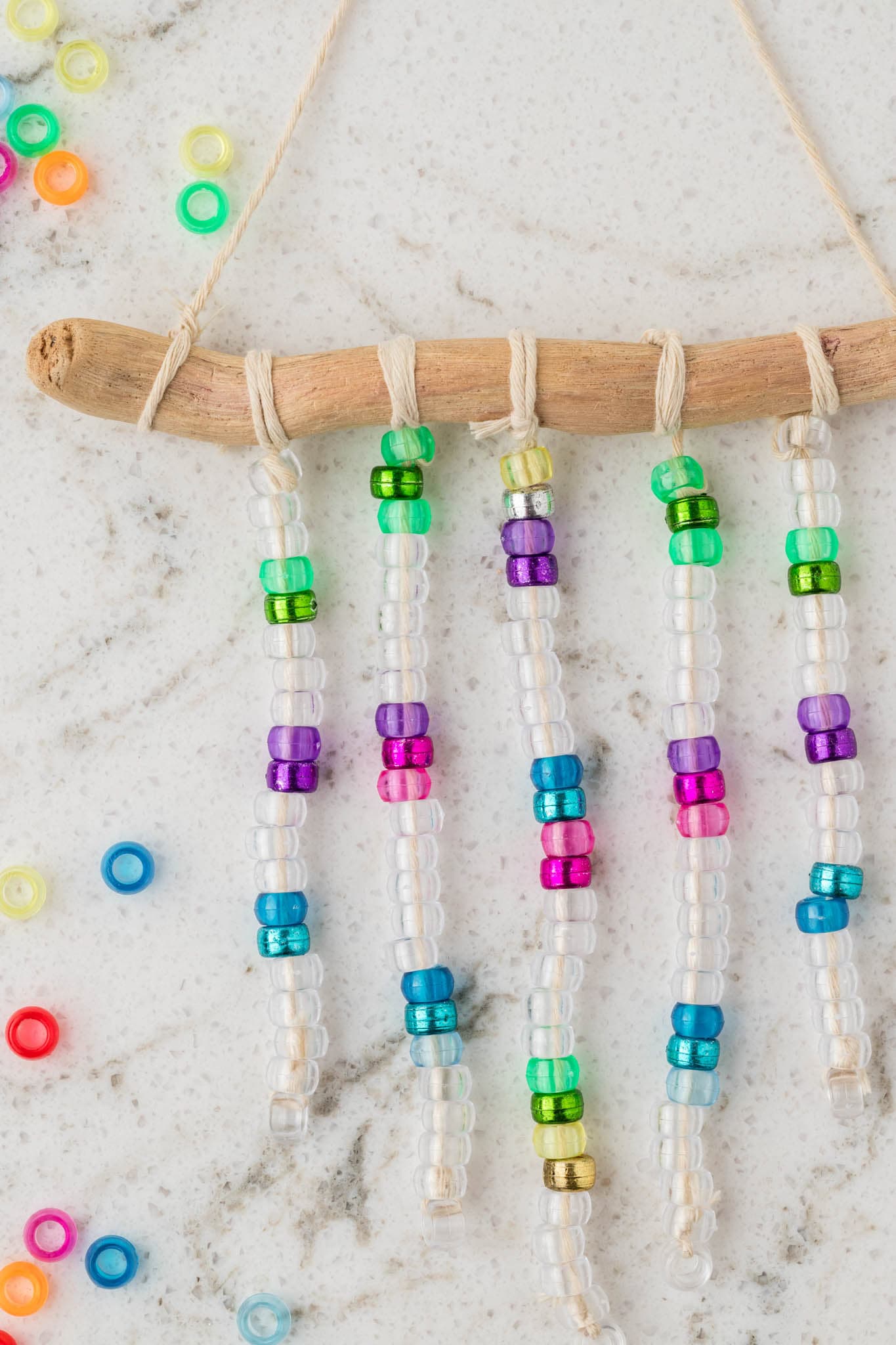 A close-up of a handmade beaded suncatcher hanging from a curved piece of driftwood, with five strands of clear and colorful beads attached using twine. The beads include shades of green, purple, pink, blue, and gold. Scattered loose beads surround the suncatcher on a white marble surface with gray veining, enhancing the vibrant and creative crafting scene.