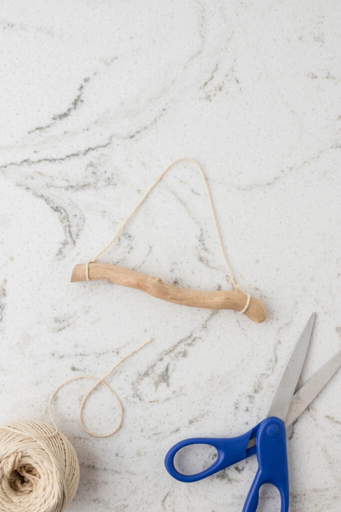 A piece of driftwood with a natural curve is tied with twine on both ends, forming a hanging loop. The setup is placed on a white marble surface with gray veining. Nearby, a spool of beige twine, a loose piece of string, and a pair of blue-handled scissors are visible, suggesting a crafting project in progress.
