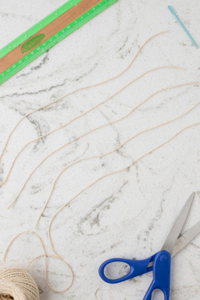 Several pieces of beige twine are laid out in parallel on a white marble surface with gray veining. A green wooden ruler, a blue plastic needle, a spool of twine, and a pair of blue-handled scissors are also present, indicating a crafting project in progress.