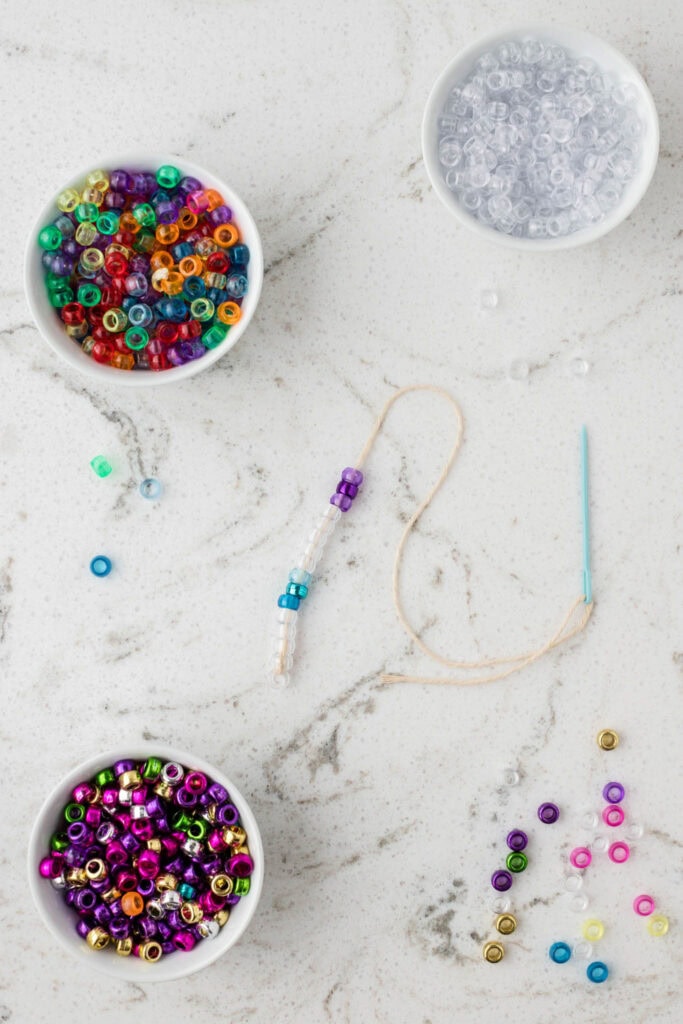 A crafting setup on a white marble surface with gray veining features three small bowls filled with colorful plastic beads—one with multicolored transparent beads, one with clear beads, and one with metallic beads. A piece of twine threaded with a mix of white, blue, and purple beads is partially completed, with a blue plastic needle attached. Scattered beads are visible around the workspace, indicating an ongoing beading project.
