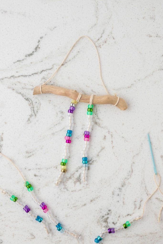 A handmade beaded suncatcher in progress is displayed on a white marble surface with gray veining. A curved driftwood piece serves as the base, with two strands of clear and colorful beads attached to it using twine. Additional beaded strands are being assembled nearby, along with a blue plastic needle and extra twine, indicating an ongoing craft project.