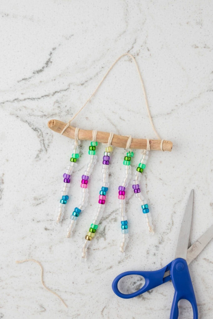 A completed beaded suncatcher hangs from a curved piece of driftwood, with five strands of clear and colorful beads attached using twine. The suncatcher is displayed on a white marble surface with gray veining. A pair of blue-handled scissors and a loose piece of twine are nearby, indicating the completion of the craft project.