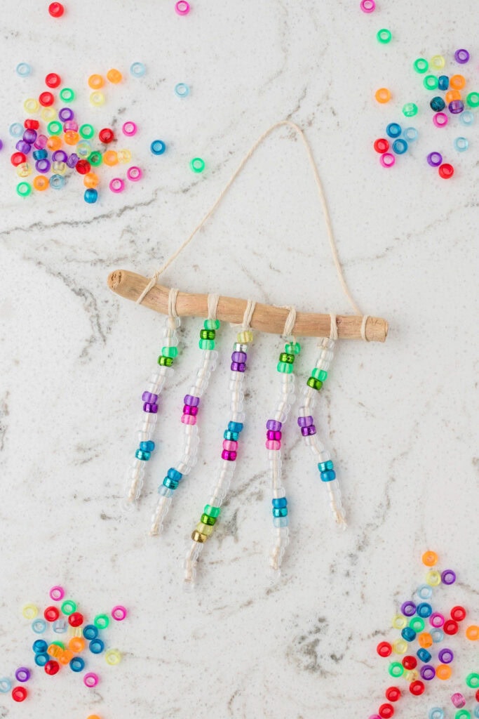 A completed beaded suncatcher hangs from a curved piece of driftwood, with five strands of clear and colorful beads attached using twine. The suncatcher is displayed on a white marble surface with gray veining. Brightly colored beads in shades of red, blue, green, yellow, purple, and orange are scattered around the workspace, enhancing the vibrant and creative crafting scene.