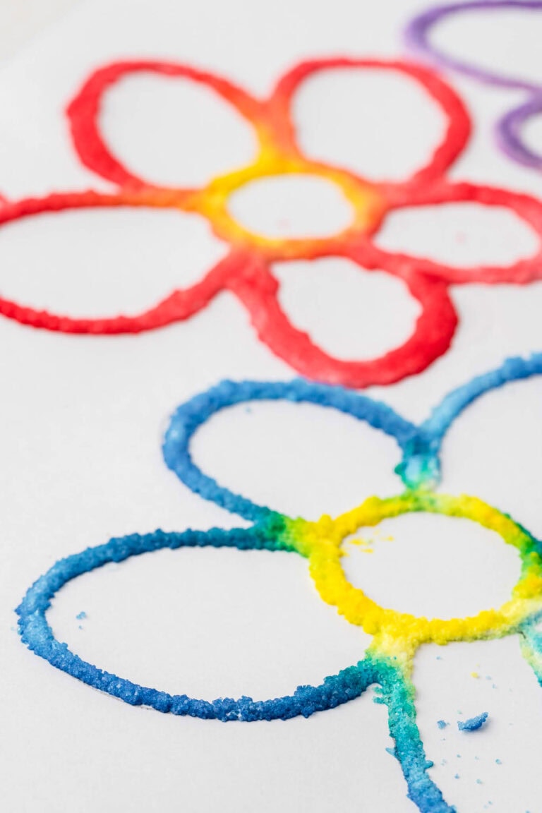 A close-up of a colorful salt painting featuring two flowers outlined in vibrant hues. The top flower has red and yellow petals, while the bottom flower transitions from blue to green and yellow. The textured salt design creates a raised, tactile effect on a white background.