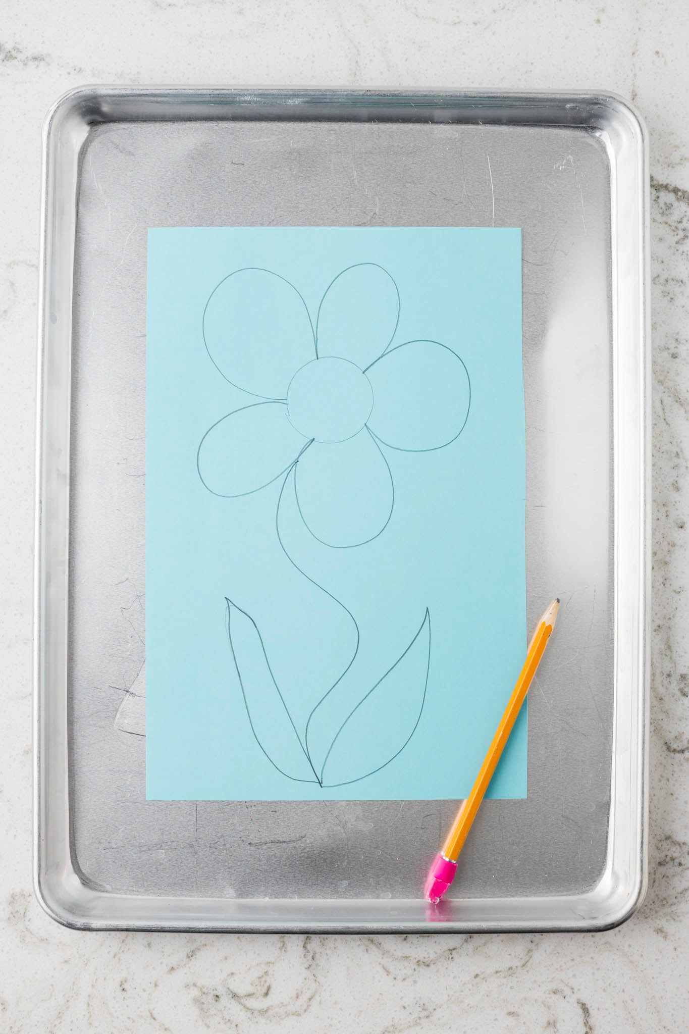 A simple pencil sketch of a flower with a stem and leaves on a blue sheet of paper, placed on a metal baking tray with a yellow pencil beside it.