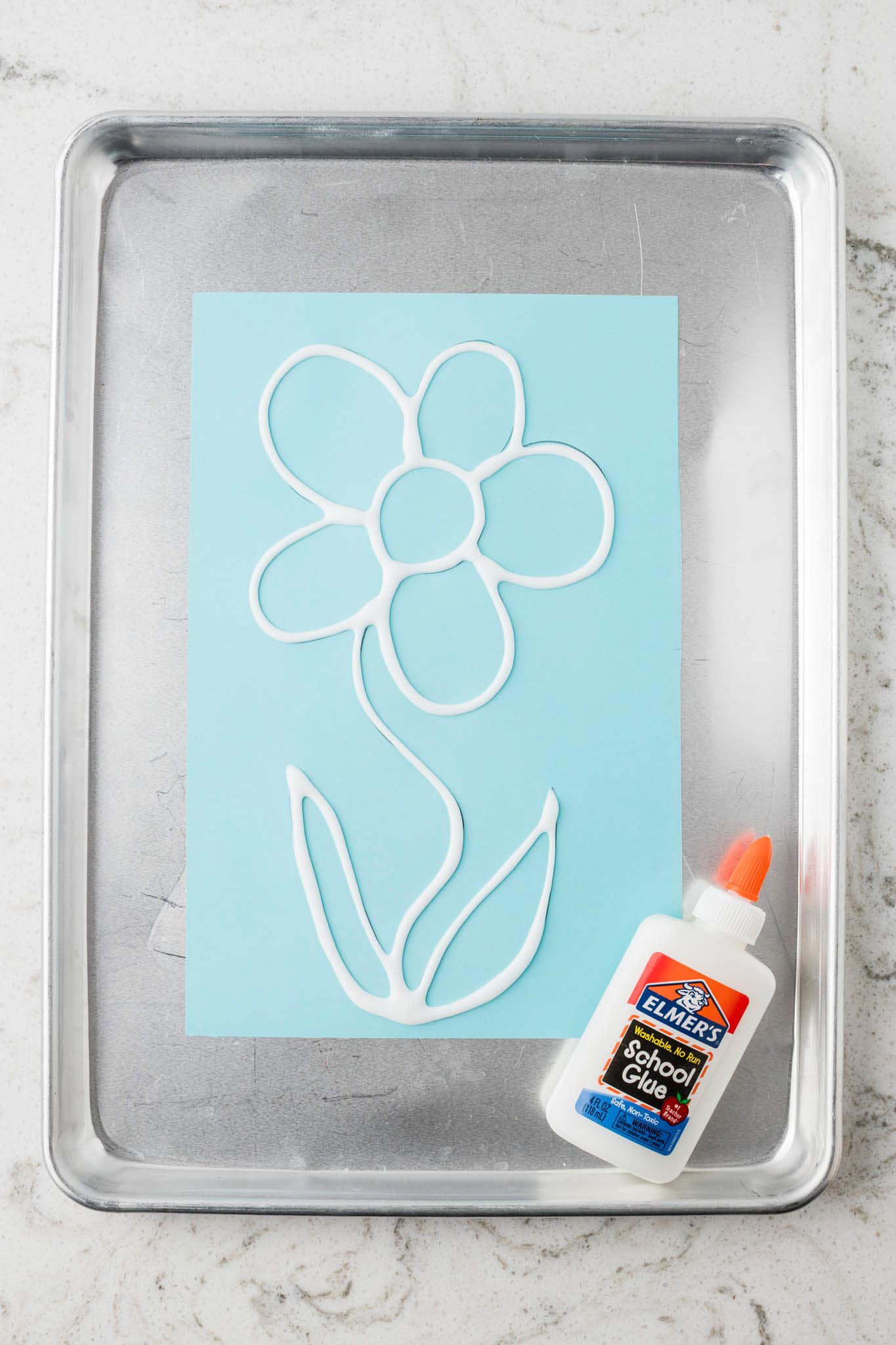 A bottle of Elmer’s glue next to the blue paper, with glue traced over the flower drawing to create raised outlines.