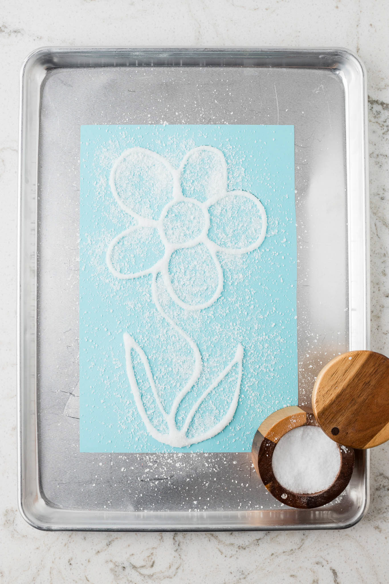 Salt sprinkled over the glued flower outline, covering the design while excess salt is visible on the tray.