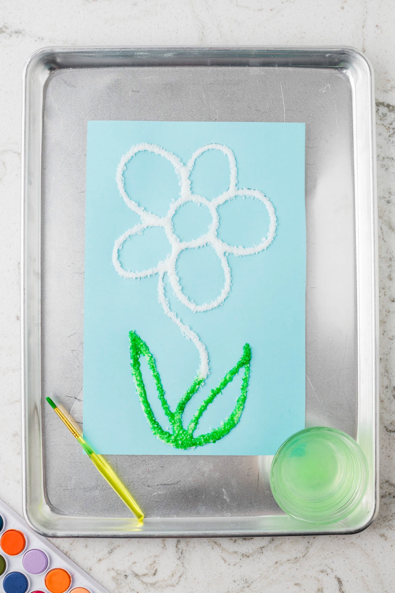 The salt painting process has begun, with green watercolor paint applied to the stem and leaves, and a paintbrush and watercolor palette nearby.