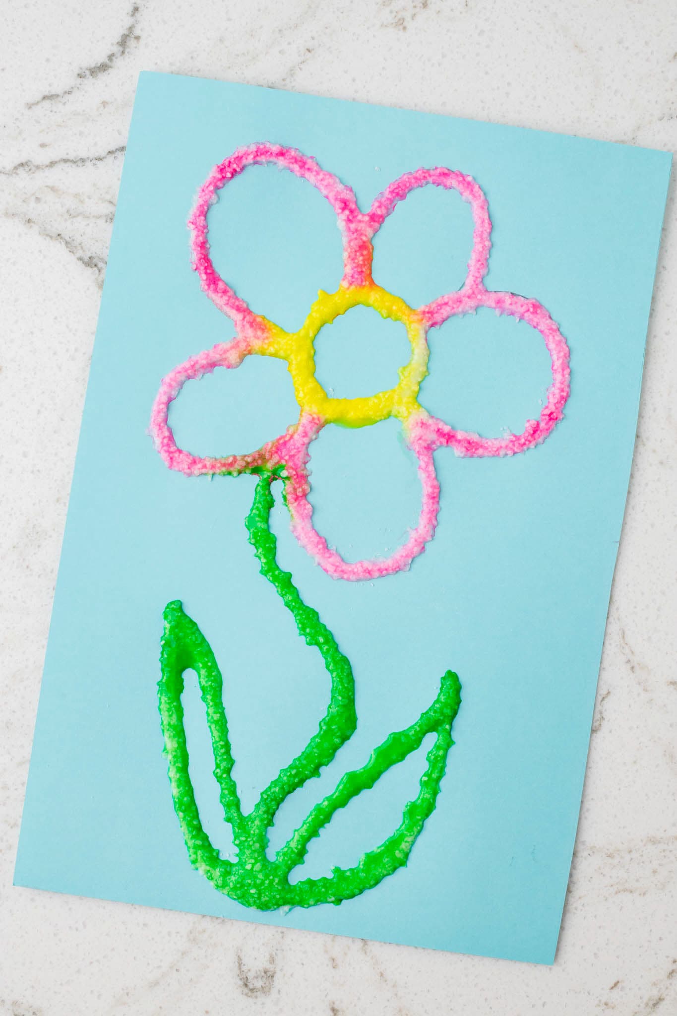 A close-up of the finished salt painting, highlighting the textured, vibrant colors of the flower on the blue paper.