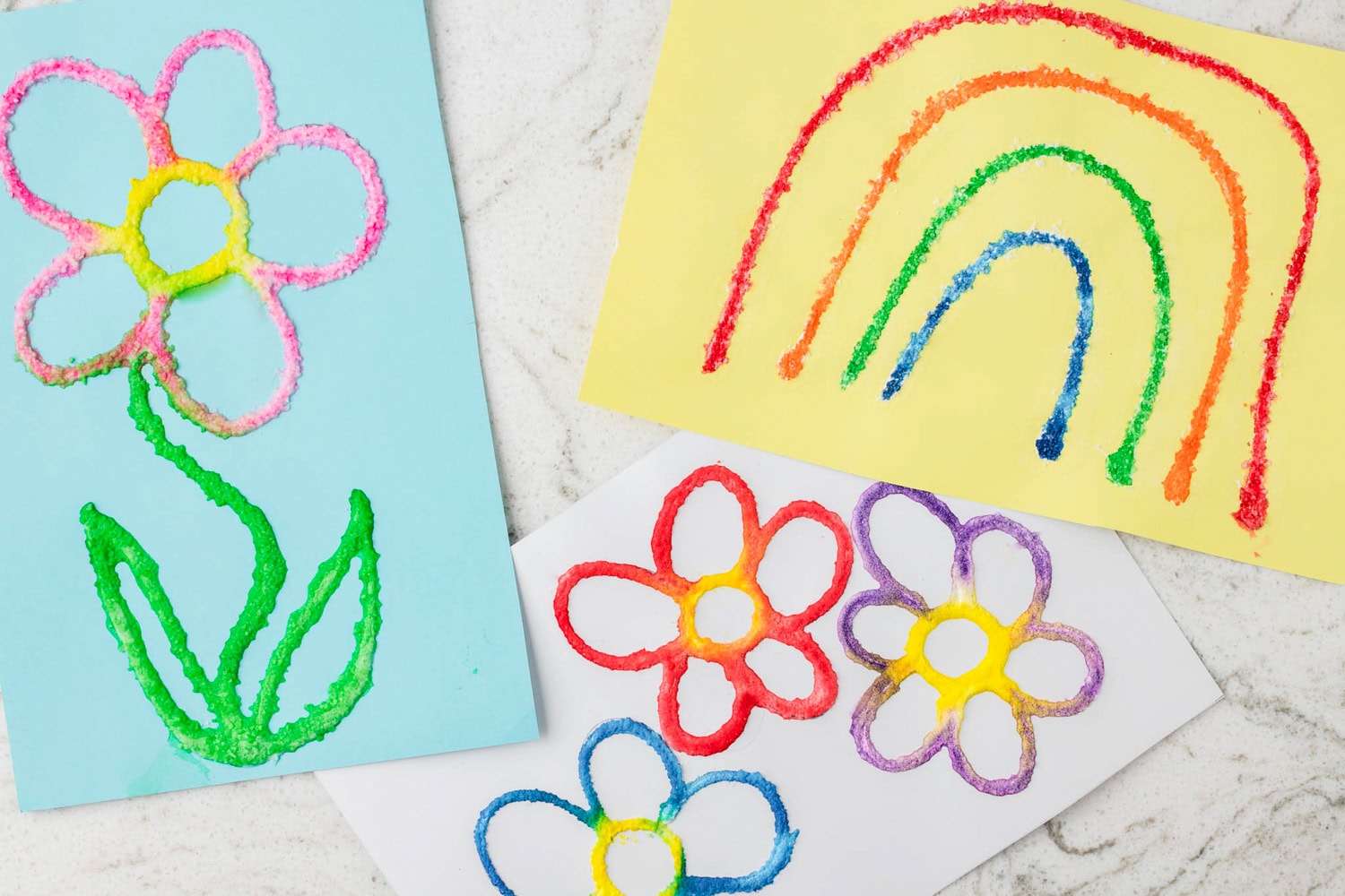 A collection of colorful salt paintings featuring a variety of designs on different colored papers. One artwork on blue paper showcases a vibrant flower with green leaves, while another on yellow paper depicts a multicolored rainbow. A third piece on white paper displays multiple flowers in red, purple, blue, and yellow. The raised salt texture enhances the designs, creating a unique tactile effect.