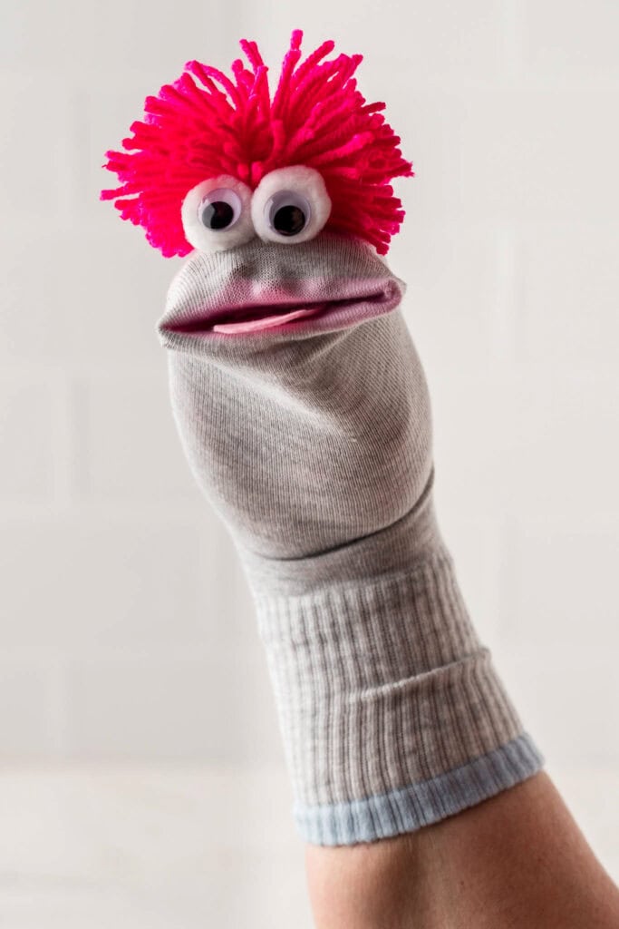 A close-up of a handmade sock puppet with a gray sock base, featuring large googly eyes, a pink felt mouth, and bright pink yarn hair. The puppet is worn on a person's hand, with the sock extending down the wrist. The background is blurred with neutral tones, making the puppet the focal point of the image.