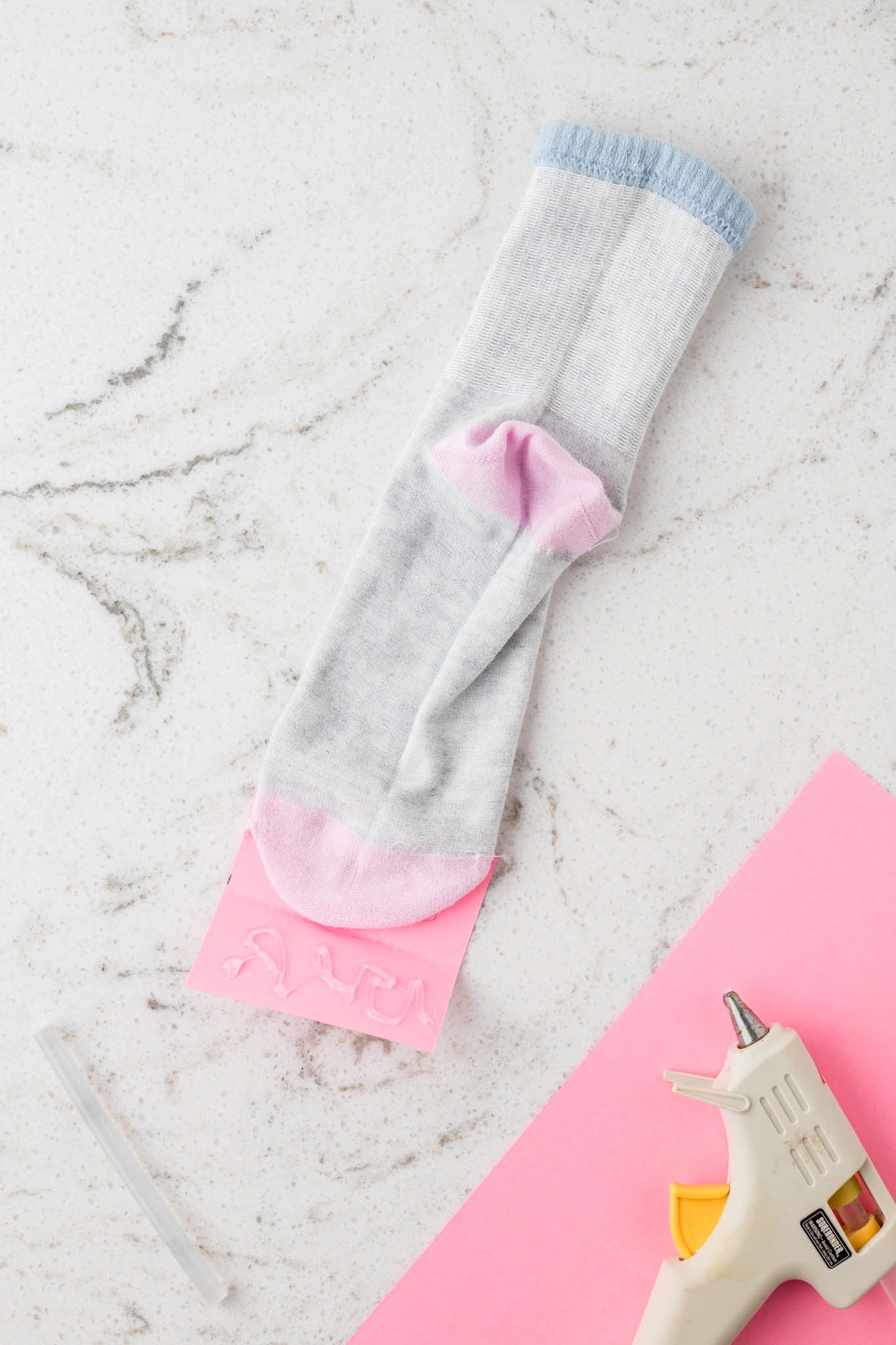 A gray sock with pink and blue accents is laid out on a white marble surface, with its toe section placed over a piece of pink felt. Hot glue is applied to the felt, likely for crafting a sock puppet. A hot glue gun and an extra glue stick are also visible nearby, suggesting an ongoing DIY project.
