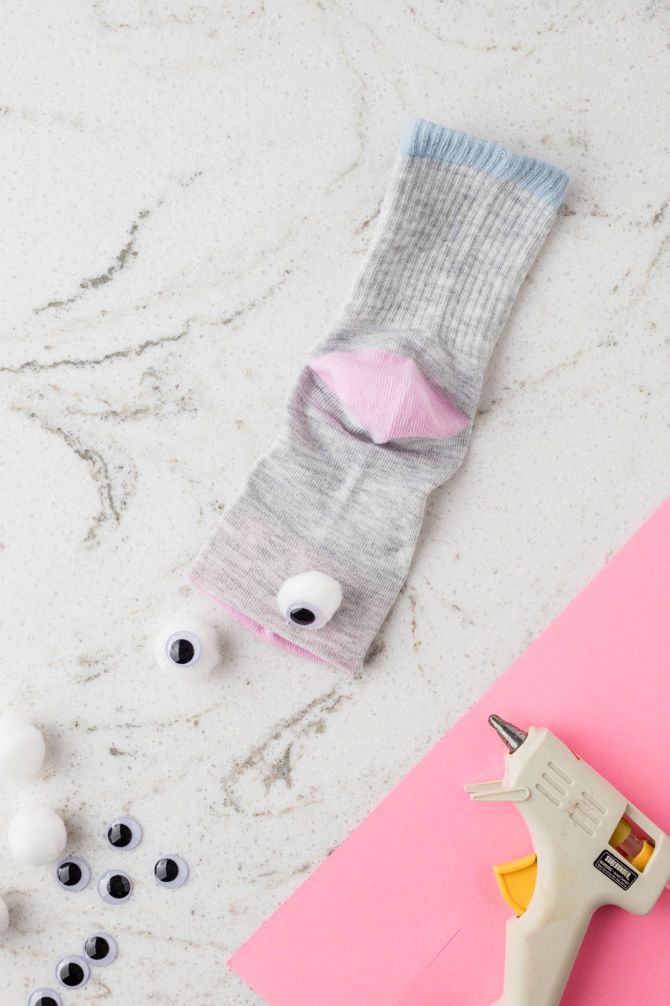 A gray sock with a blue cuff is laid out on a white marble surface, with a pink felt piece partially inserted to form the puppet's mouth. Two large googly eyes, attached to white pom-poms, are positioned near the sock. Additional crafting supplies, including more googly eyes, white pom-poms, a hot glue gun, and a sheet of pink felt, are scattered around, indicating an ongoing DIY sock puppet project.