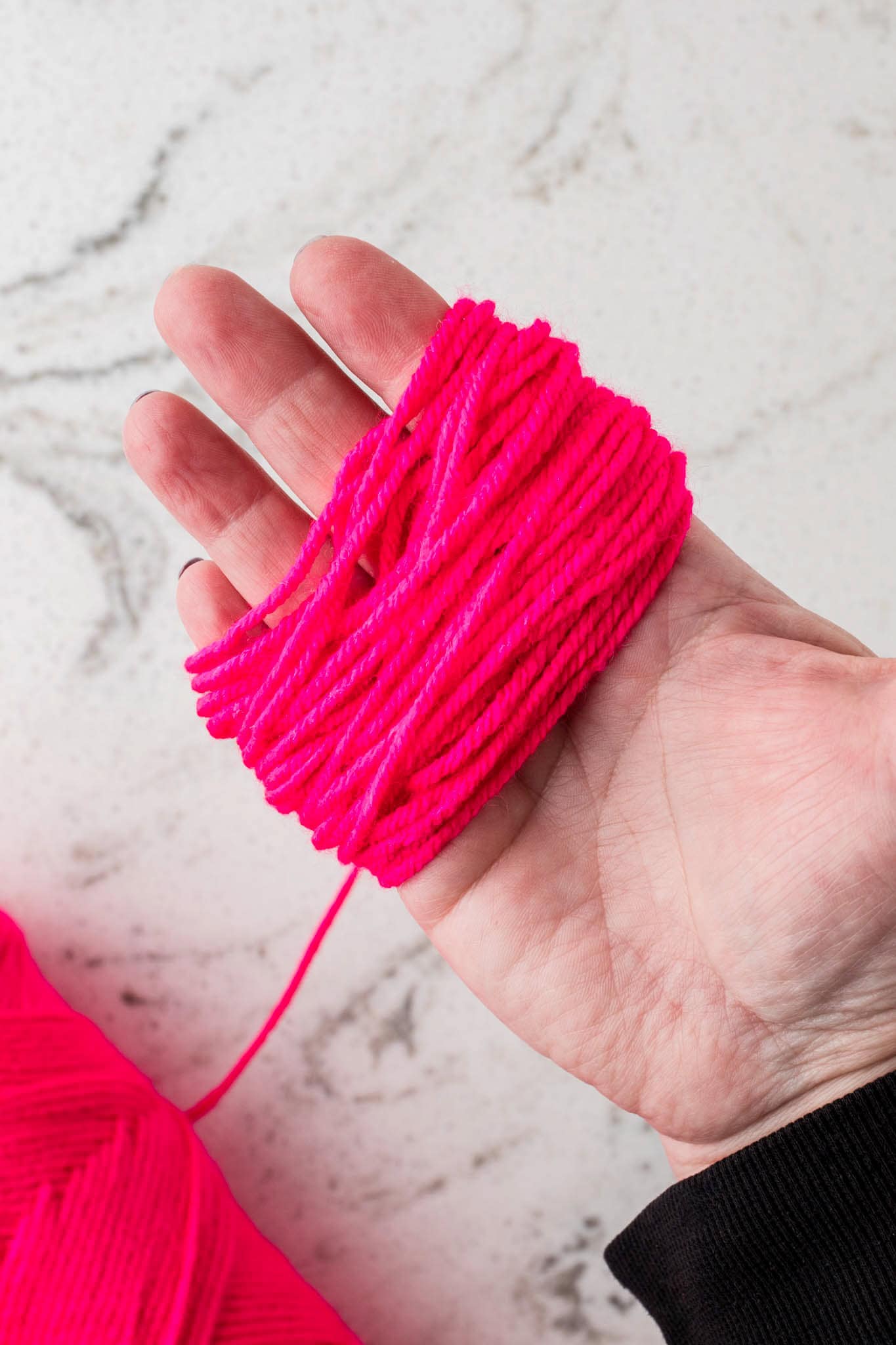 A close-up of a person's hand holding bright pink yarn wrapped around their fingers. The yarn is neatly looped multiple times, likely in preparation for crafting, such as making hair for a sock puppet. A skein of the same pink yarn is visible in the background on a white marble surface. The person is wearing a black long-sleeve garment, which contrasts with the vibrant color of the yarn.
