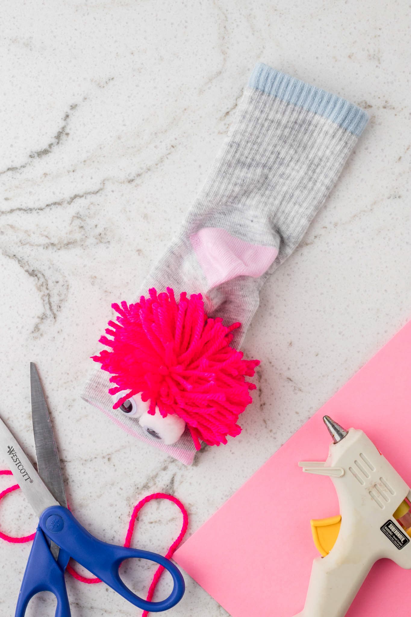 A gray sock puppet in progress is laid out on a white marble surface. It has large googly eyes attached to white pom-poms and a bright pink yarn tuft for hair. The pink felt inside the sock forms the puppet’s mouth. Surrounding the sock are crafting tools, including a pair of blue-handled scissors, a hot glue gun, a piece of pink felt, and a loose strand of pink yarn, indicating an ongoing DIY project.