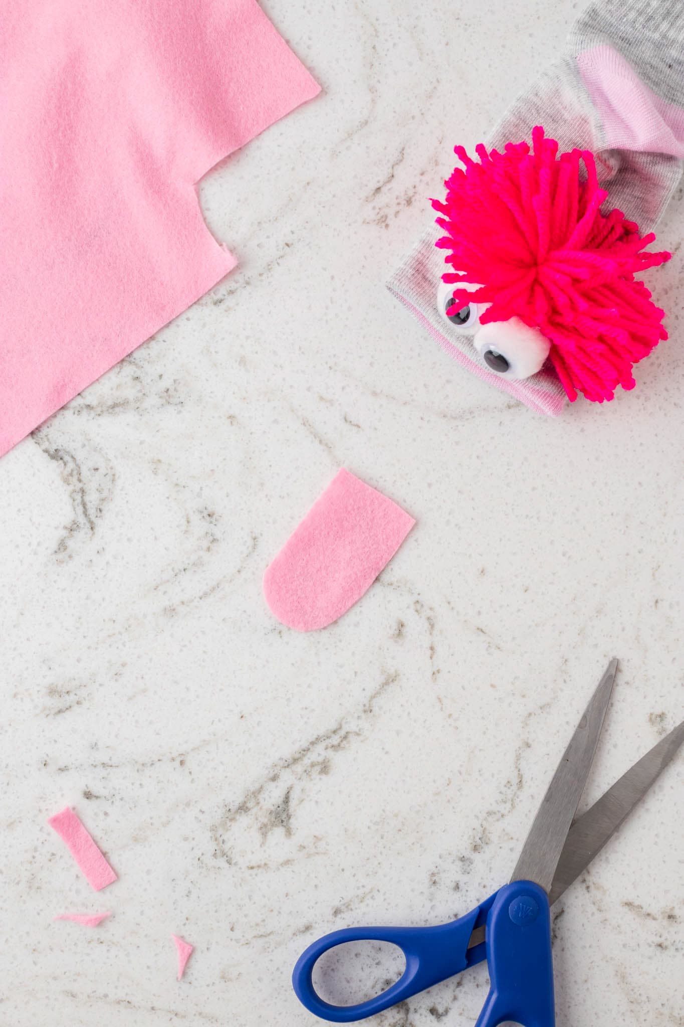 A gray sock puppet with bright pink yarn hair and googly eyes is placed on a white marble surface. Nearby, a piece of pink felt with a rounded edge is cut out, likely for the puppet’s tongue. Additional crafting materials, including a larger piece of pink felt, small felt scraps, and a pair of blue-handled scissors, are scattered around, indicating an ongoing DIY project.
