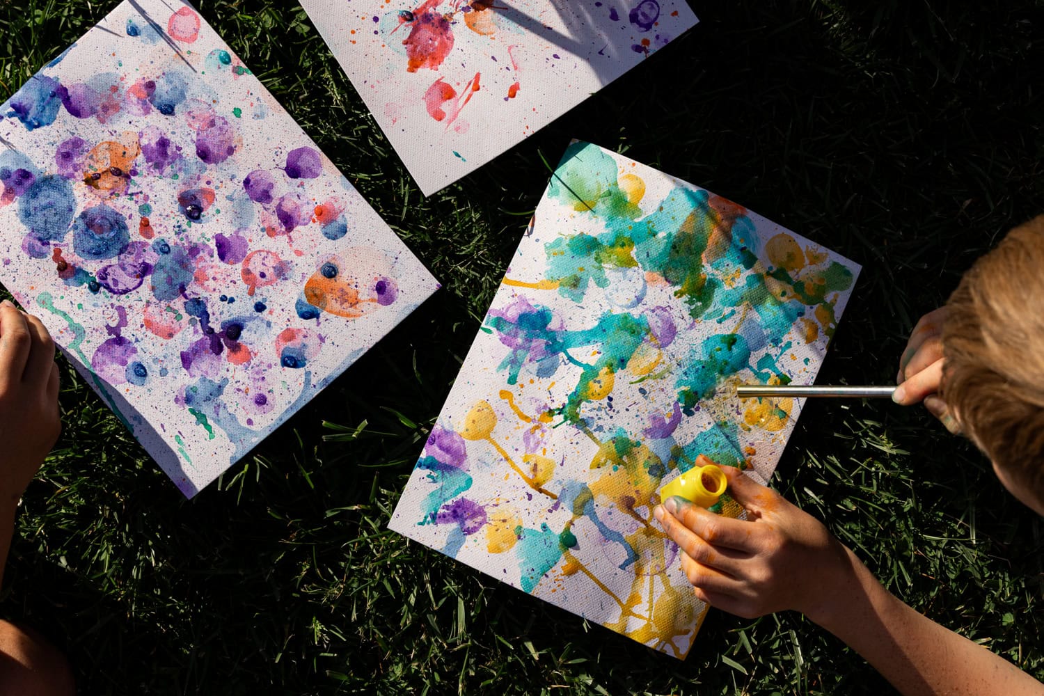 Children’s hands painting on white paper with vibrant watercolor splatters in shades of purple, orange, green, and blue, outdoors on green grass.