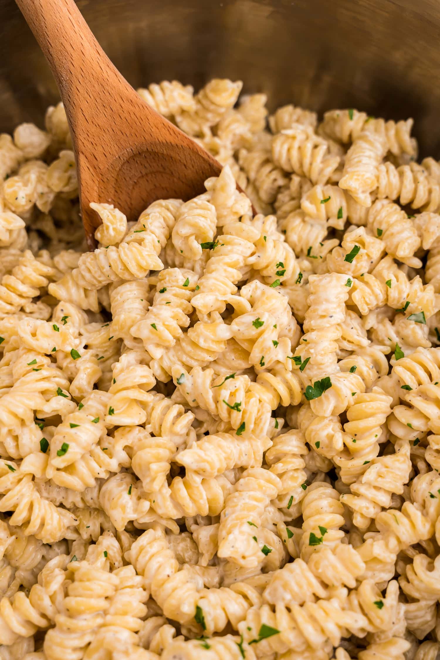 Creamy rotini pasta in a pot being stirred with a wooden spoon, garnished with finely chopped parsley for a touch of color and freshness.
