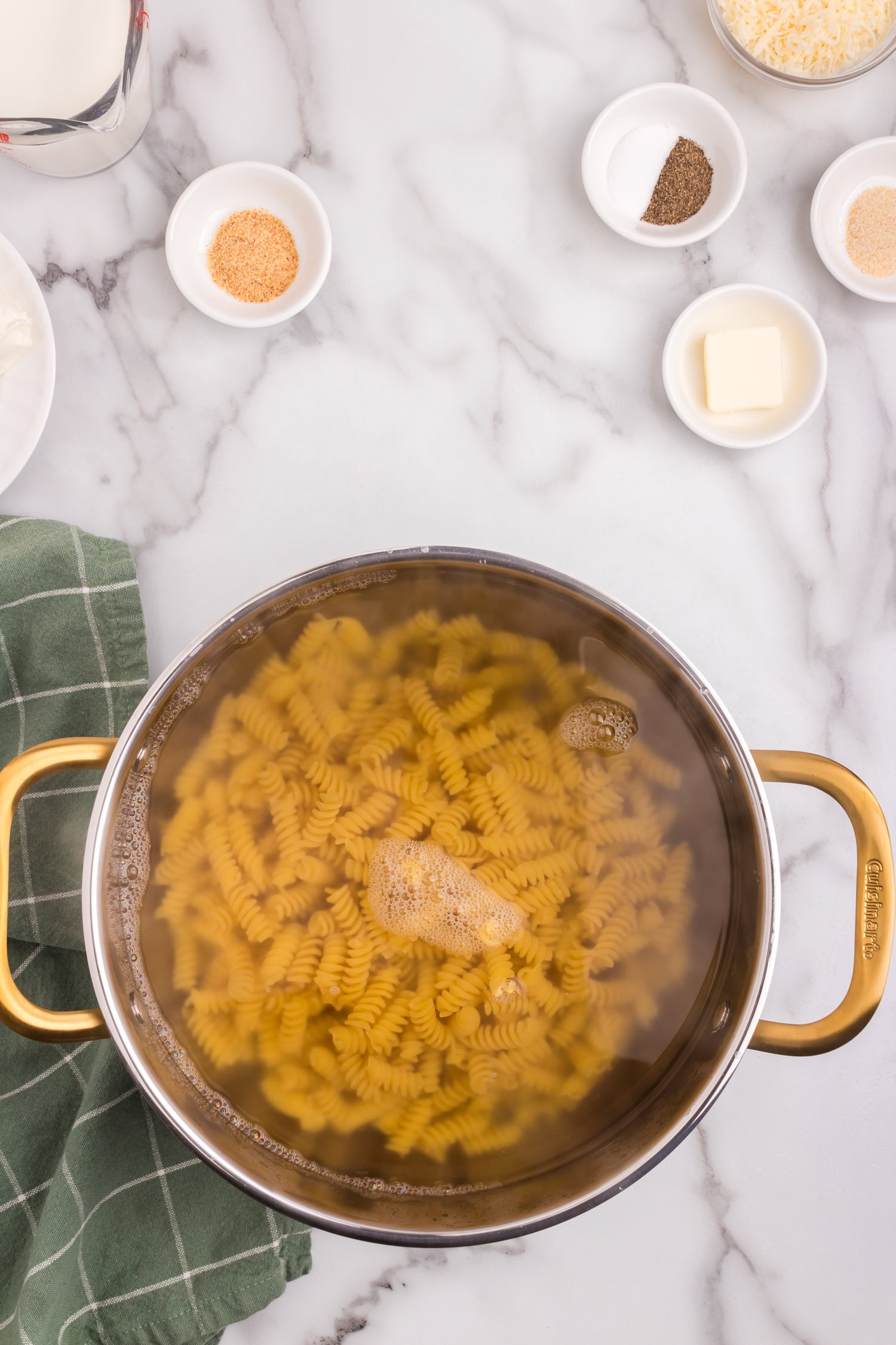 A pot filled with uncooked rotini pasta, surrounded by small bowls of ingredients including butter, cream cheese, shredded cheese, and seasoning.