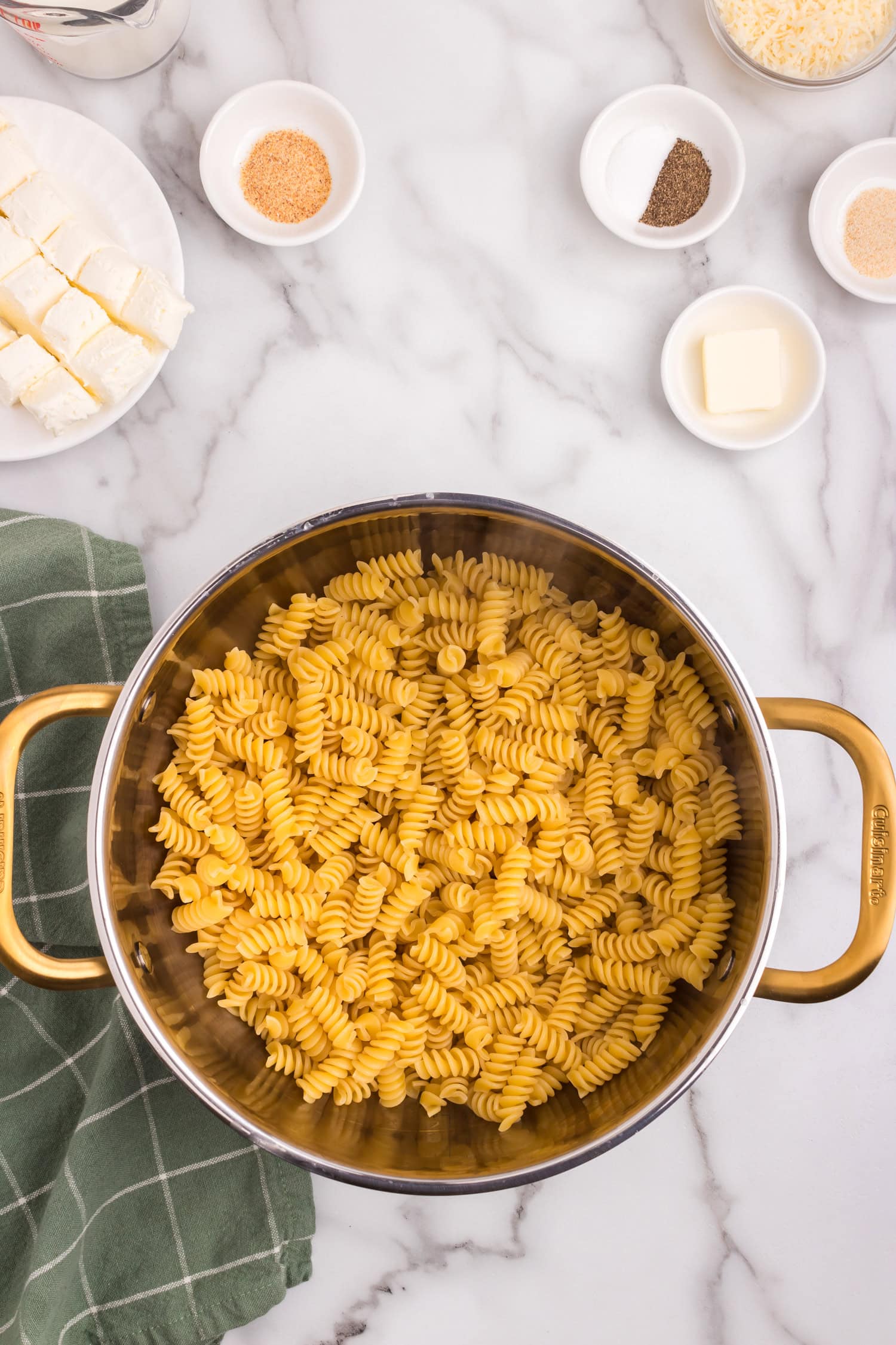 A pot filled with uncooked rotini pasta, surrounded by small bowls of ingredients including butter, cream cheese, shredded cheese, and seasoning.