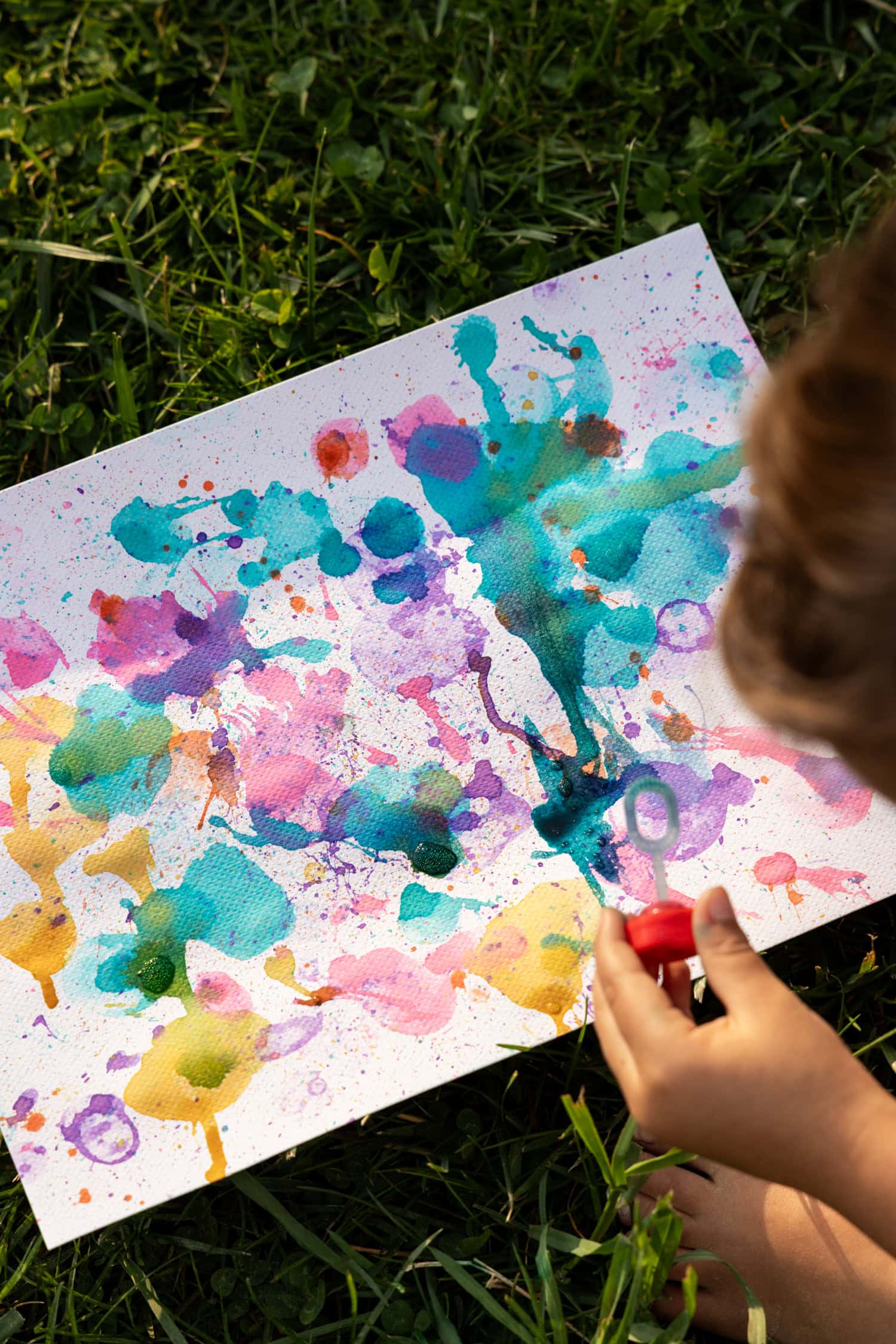 A child holding a red bubble wand over a canvas filled with colorful watercolor splashes and bubble prints on the grass.