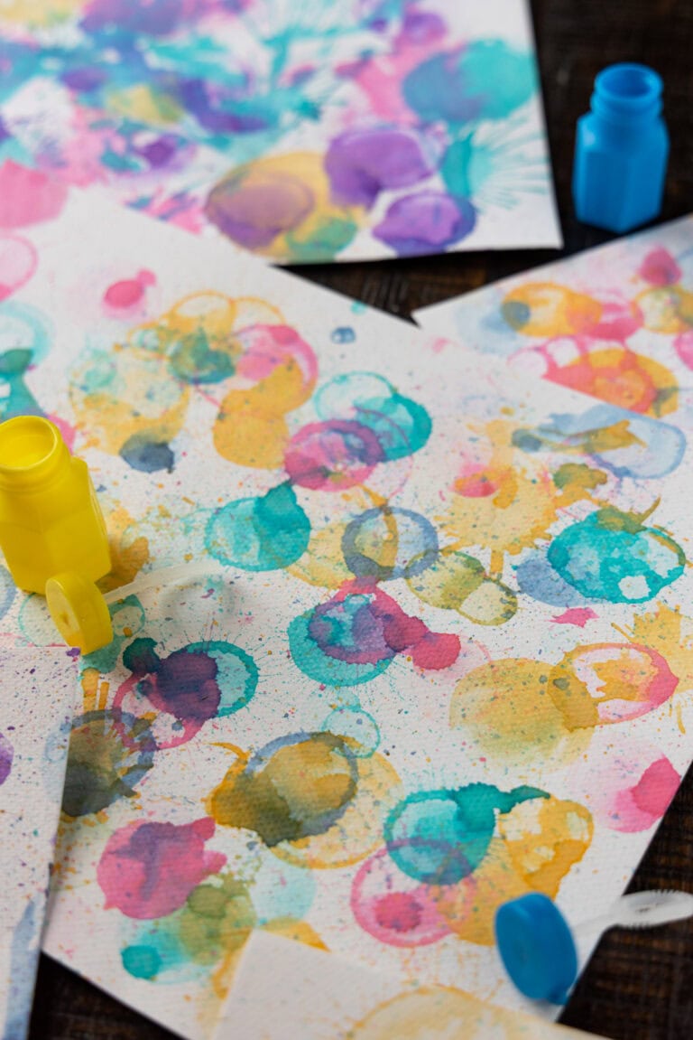 A close-up of multiple finished bubble paintings with overlapping colorful circles and splatters in vibrant tones of yellow, pink, blue, and purple, with yellow and blue bubble containers nearby.