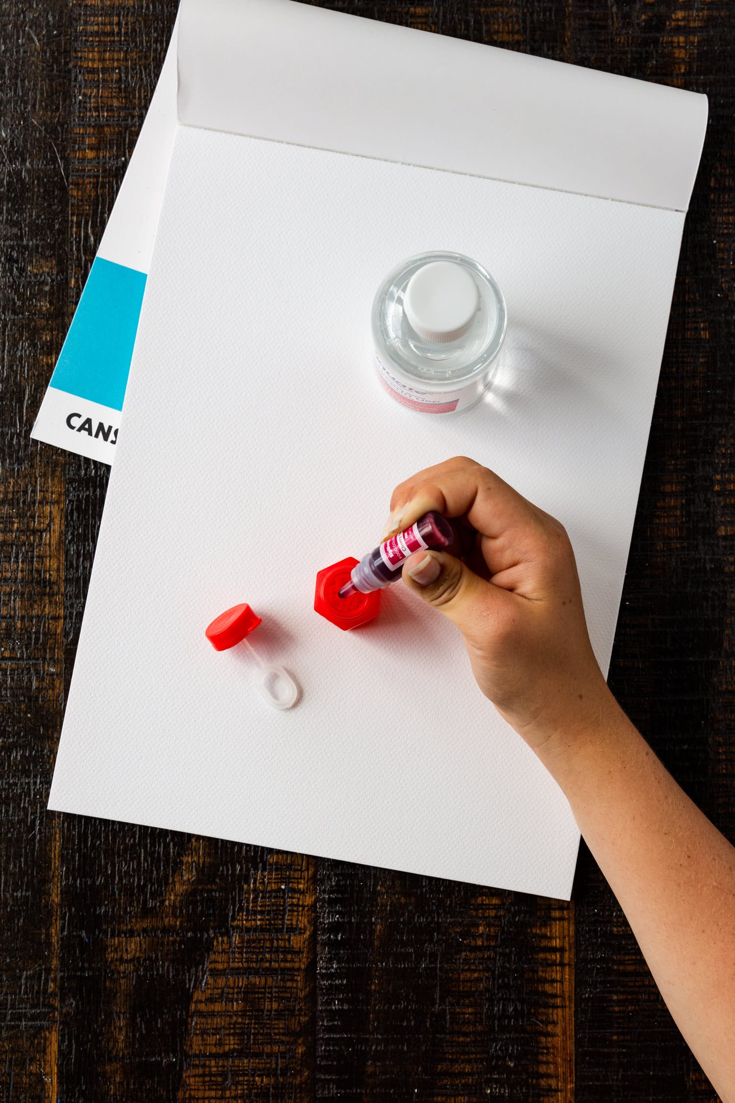 : A hand squeezing red liquid dye into a red bubble container on a blank white art pad, preparing materials for bubble painting.