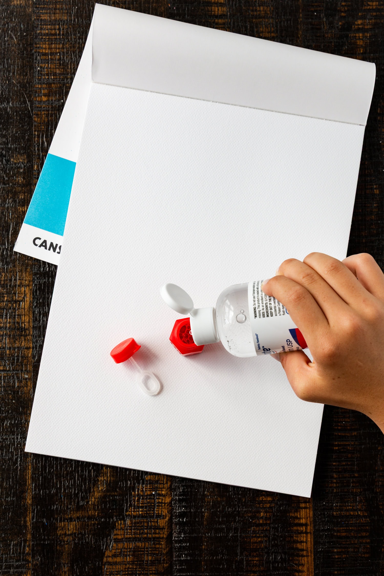 A hand pouring clear liquid, likely soap or bubble solution, into a red container on a white canvas pad.