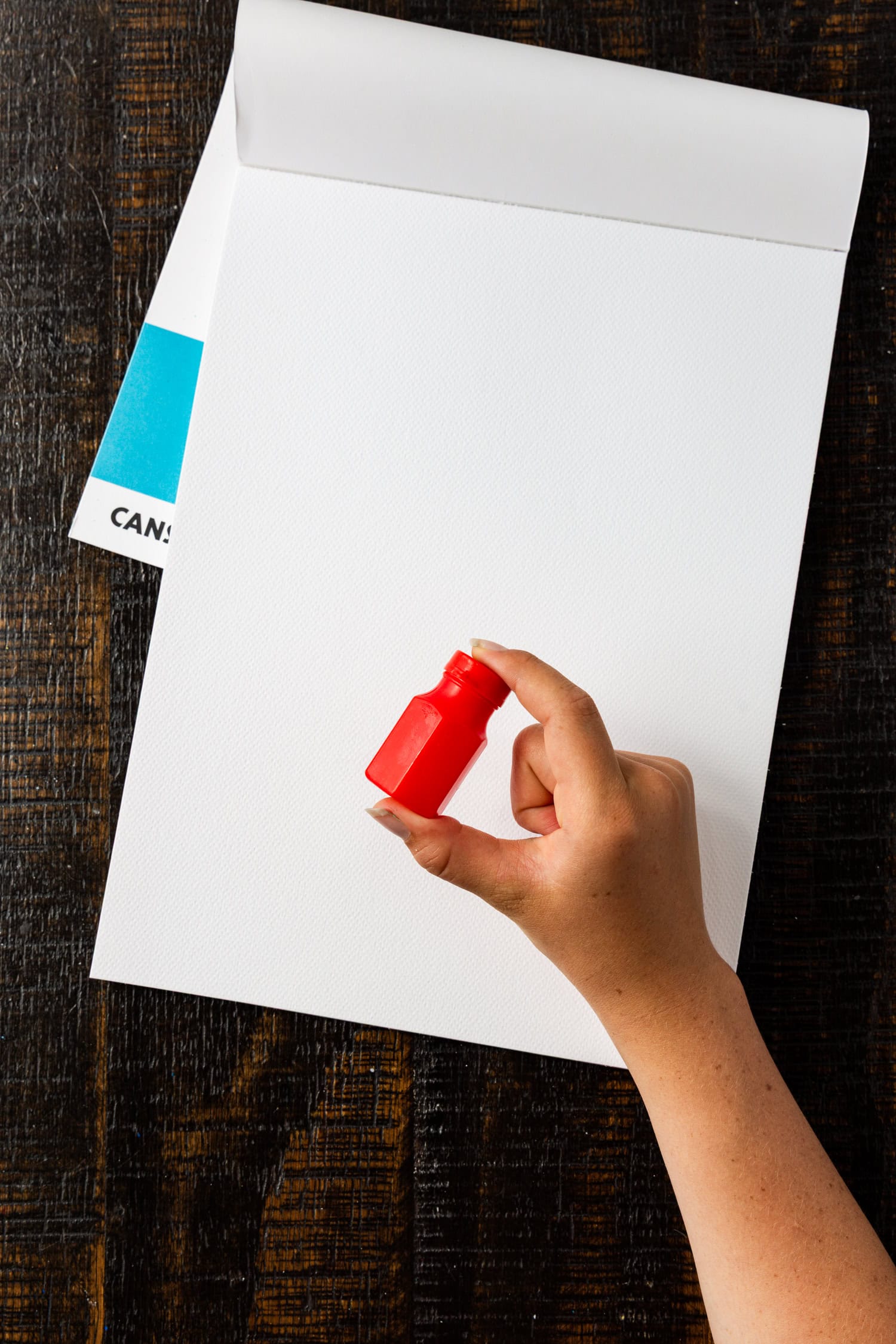 A hand holding a closed red container, likely filled with bubble solution, above a white art pad.