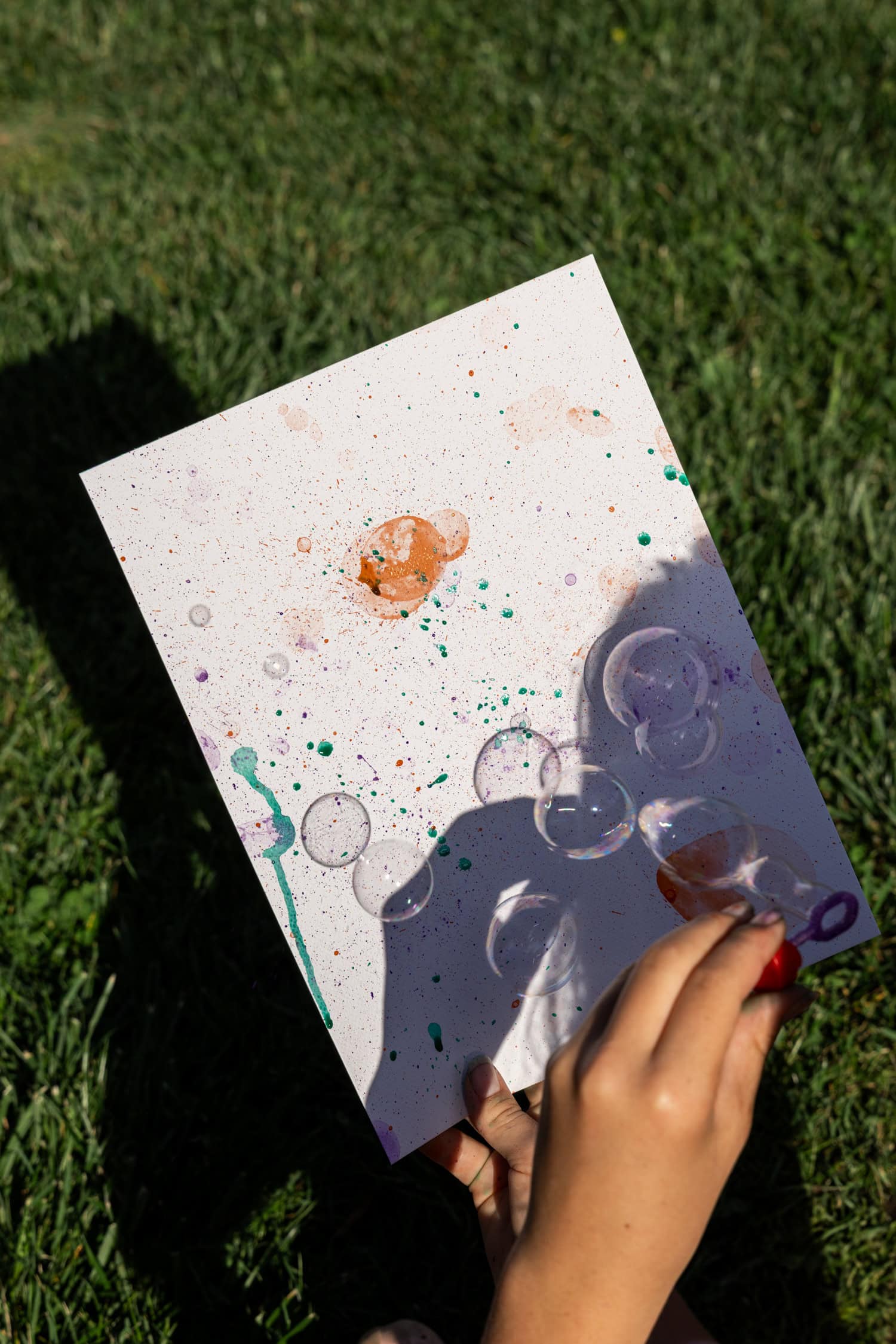 A child holding a painted sheet with colorful bubble prints and splatters while blowing bubbles outdoors on the grass.