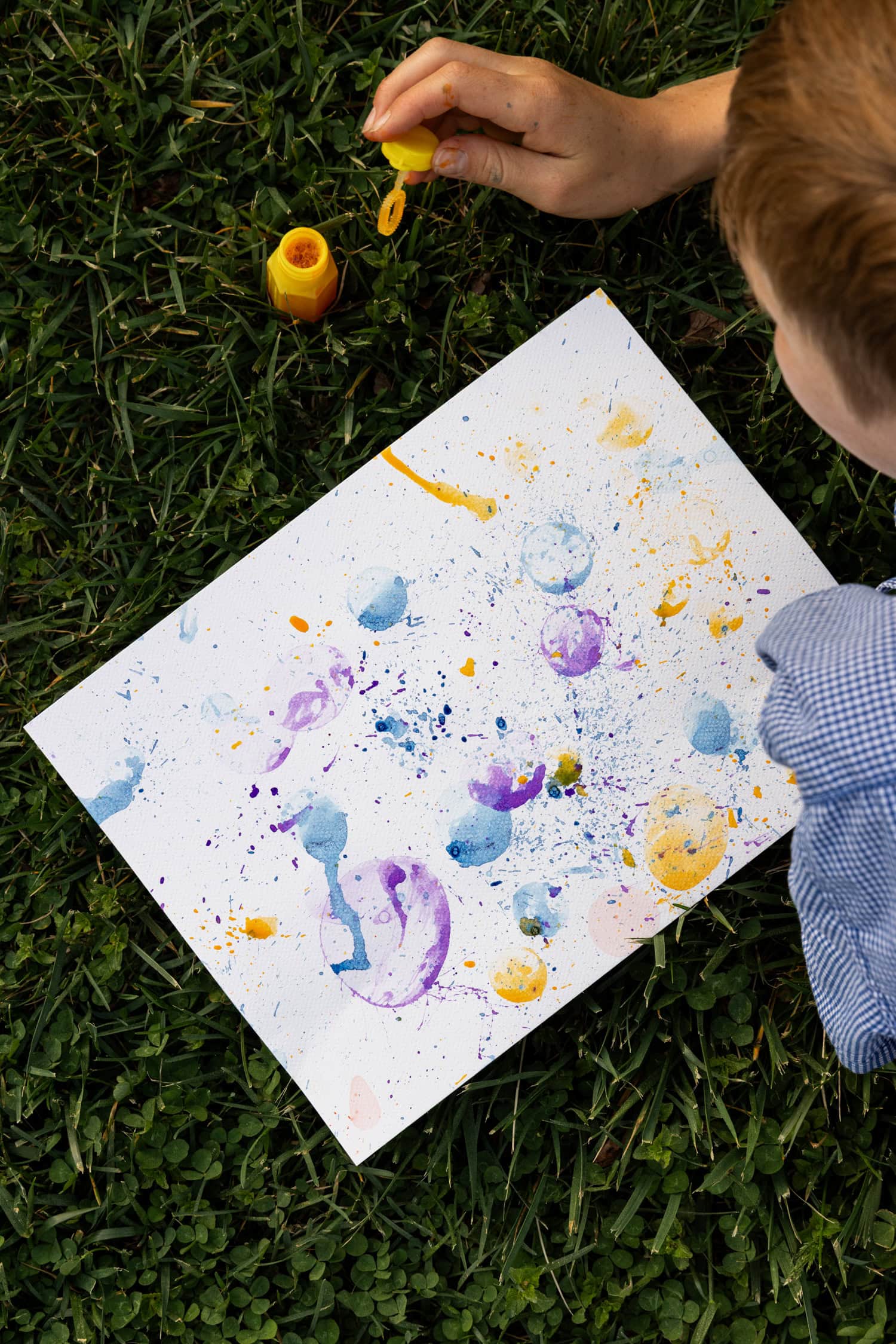 A child with a yellow bubble wand creating art with colorful paint splatters and bubble prints on a white canvas in the grass.