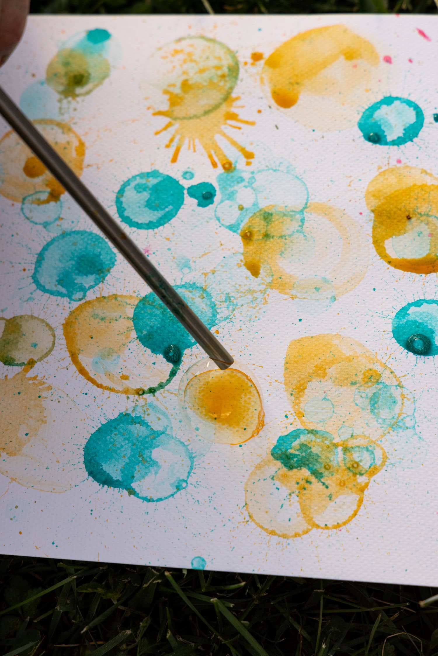 A close-up of a painting with yellow and teal bubble prints and splashes, with a metal straw-like tool pointing to a yellow bubble mark.