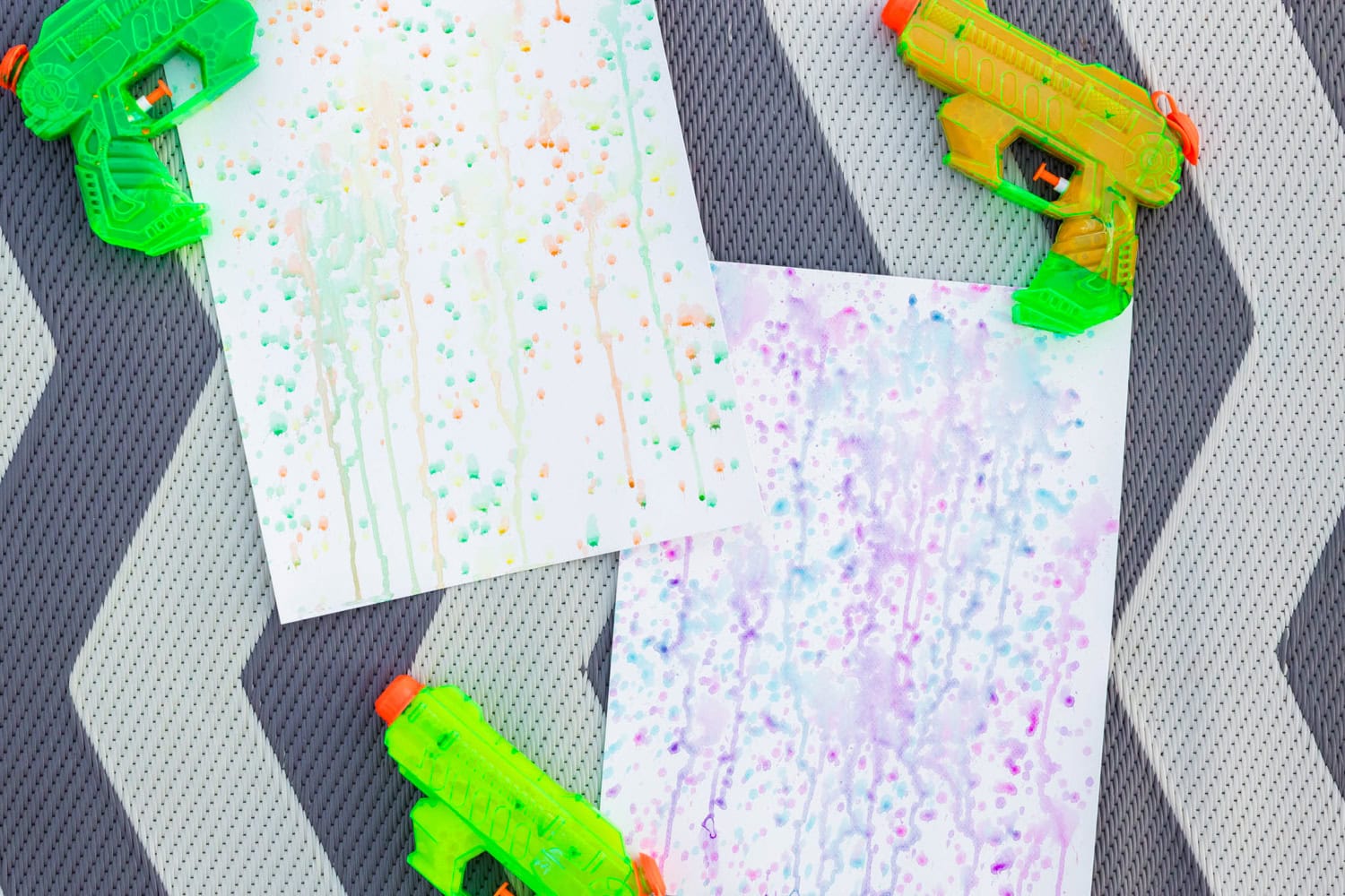 Brightly colored squirt gun paintings on paper with watercolor-like splatters and drips in green, orange, and purple tones, surrounded by neon green and orange water guns, laid out on a textured chevron-patterned mat.