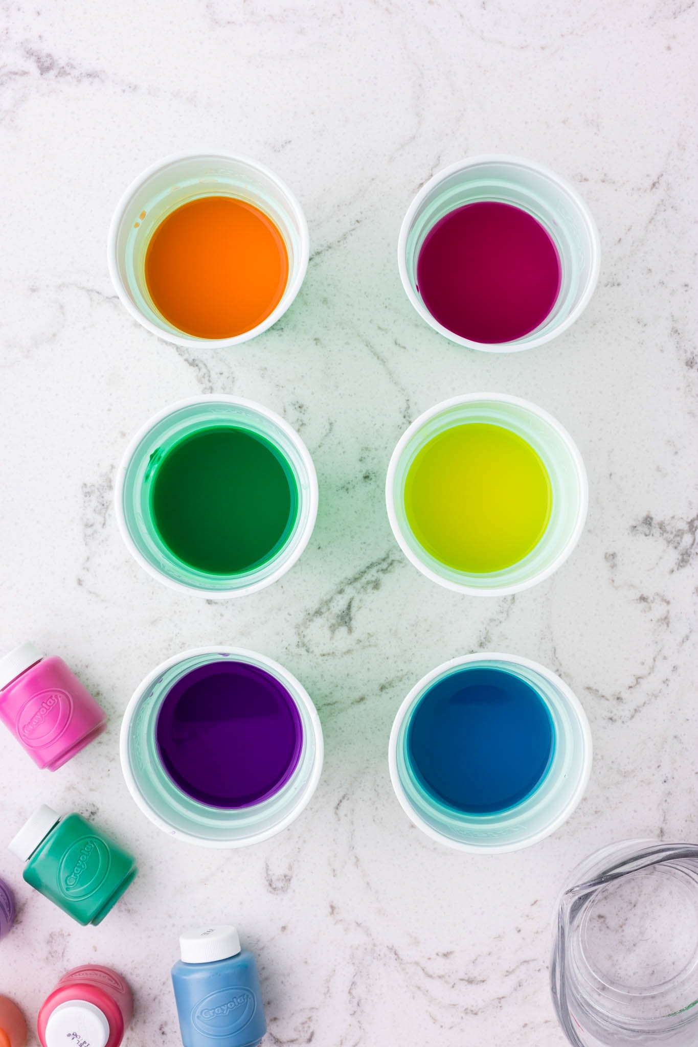 Six small white cups filled with vibrant liquid paint in orange, magenta, green, yellow, purple, and blue, arranged neatly on a white marble surface, with additional small paint bottles scattered around.