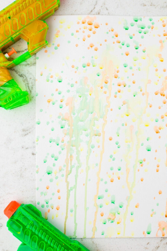 A squirt gun painting on a white surface indoors, showcasing a detailed view of colorful paint splatters and drips in green, orange, and yellow hues, with neon green water guns placed nearby.