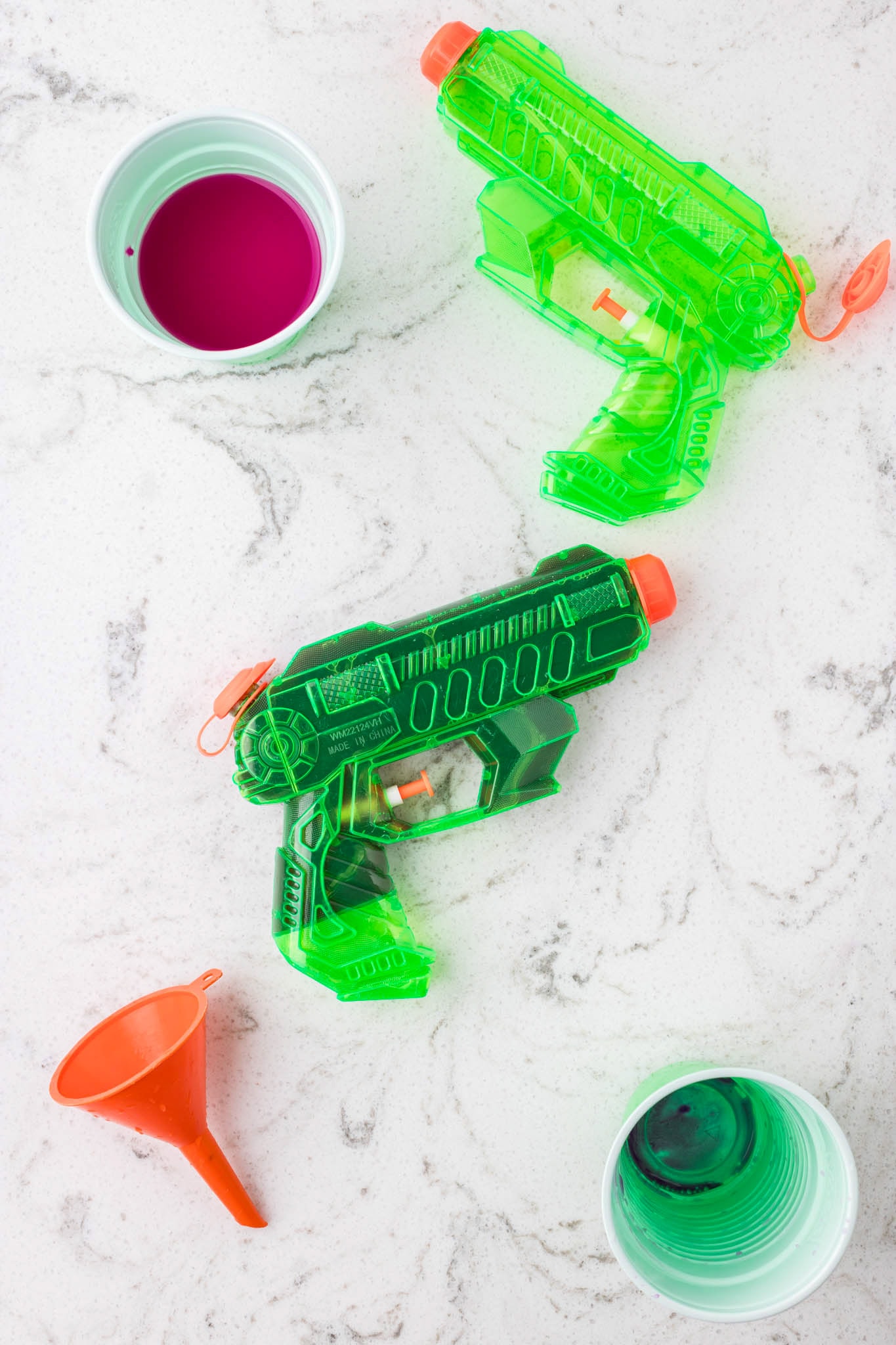 Two neon green water guns lying on a white marble surface, with a small orange funnel and cups of colorful liquid paint nearby, ready for filling.