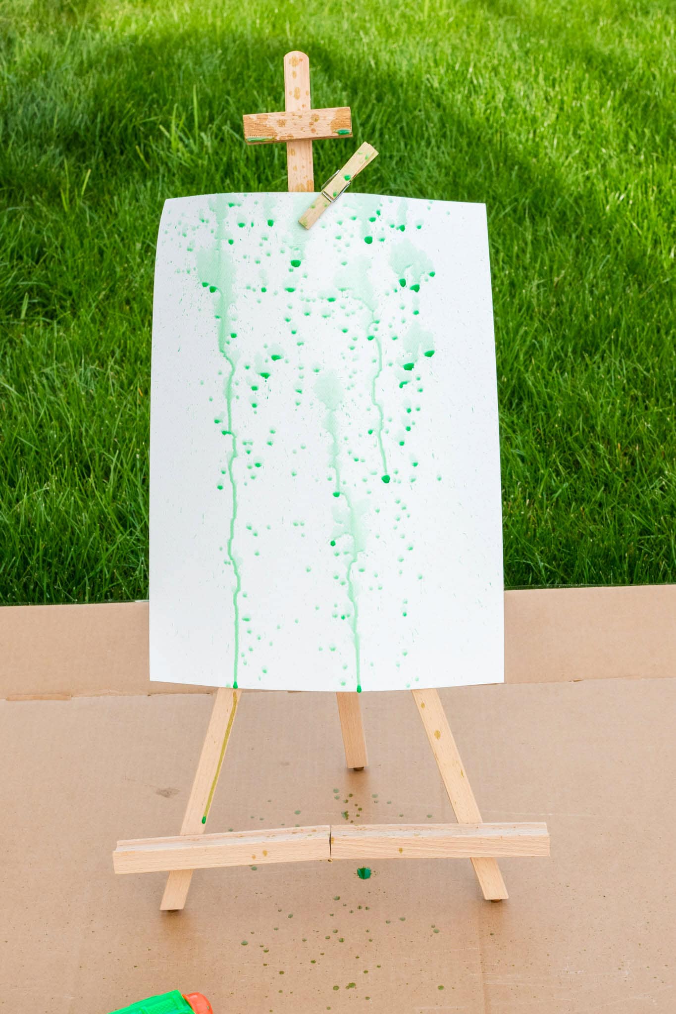 A white sheet of paper on a wooden easel outdoors, showing green paint splatters and drips created with a squirt gun, with some paint drops scattered on the cardboard base.